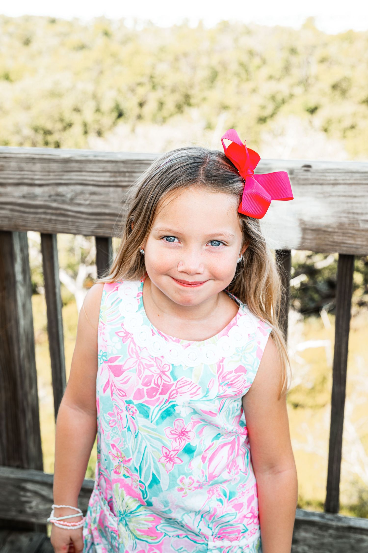 Young girl with blue eyes wearing a floral dress and red bow hair clip smiles outdoors near a wooden fence.