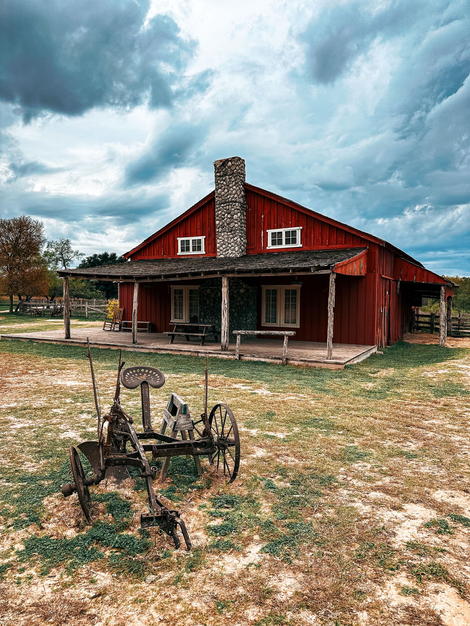 Rustic red barn with stone chimney and vintage farm equipment on dry grassland.
