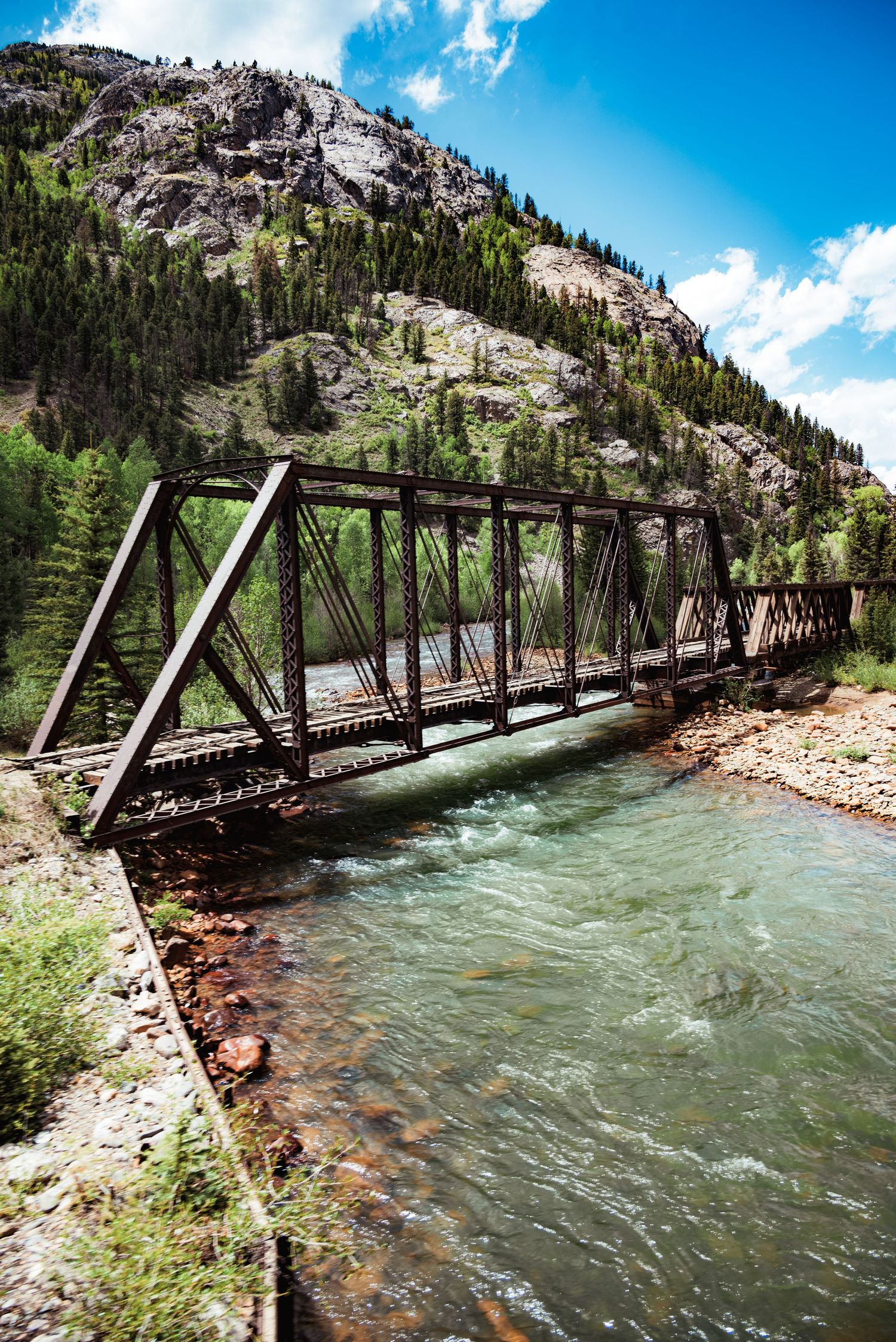 Historic iron truss railroad bridge spanning a rushing turquoise river in a mountain valley.