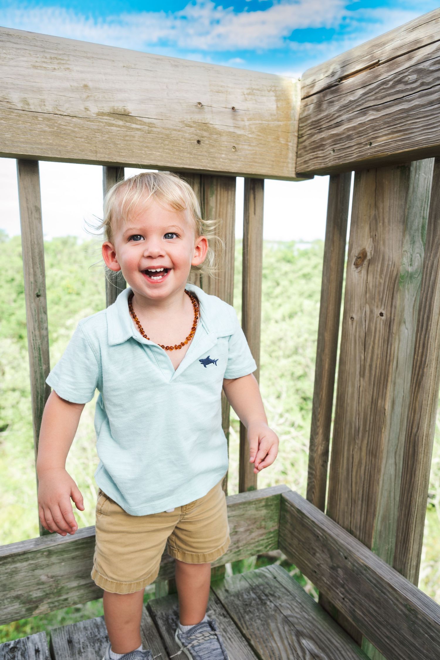 Happy toddler boy in light blue polo shirt and khaki shorts smiling on a wooden deck outdoors.