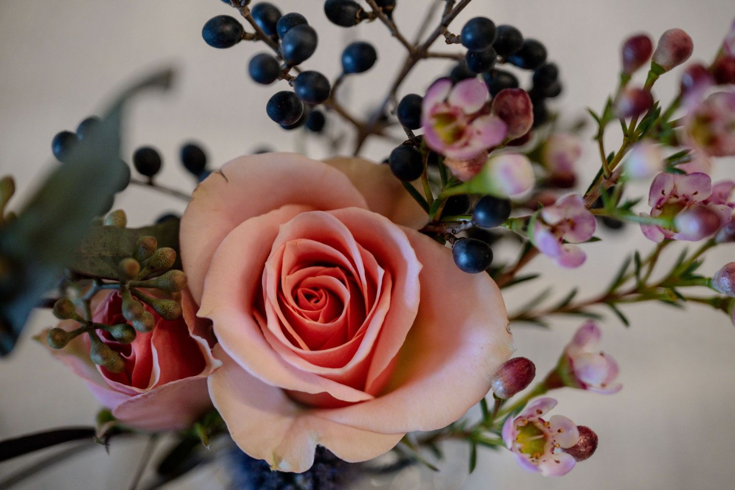 Close-up of a soft pink rose surrounded by blue berries and delicate pink wax flowers in a bouquet.