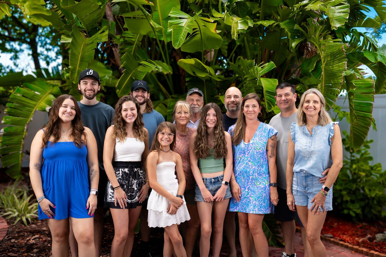 Large family group smiling together outdoors in front of tropical banana trees in a lush garden setting.