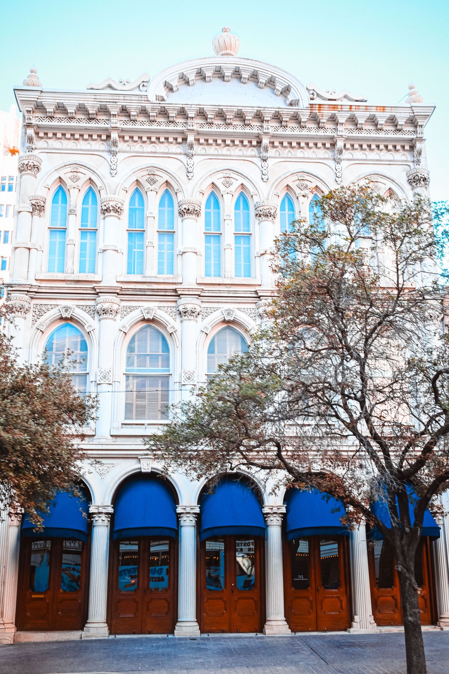 Ornate Victorian-style building facade with Gothic arched windows, blue awnings, and wooden doors under a clear sky.