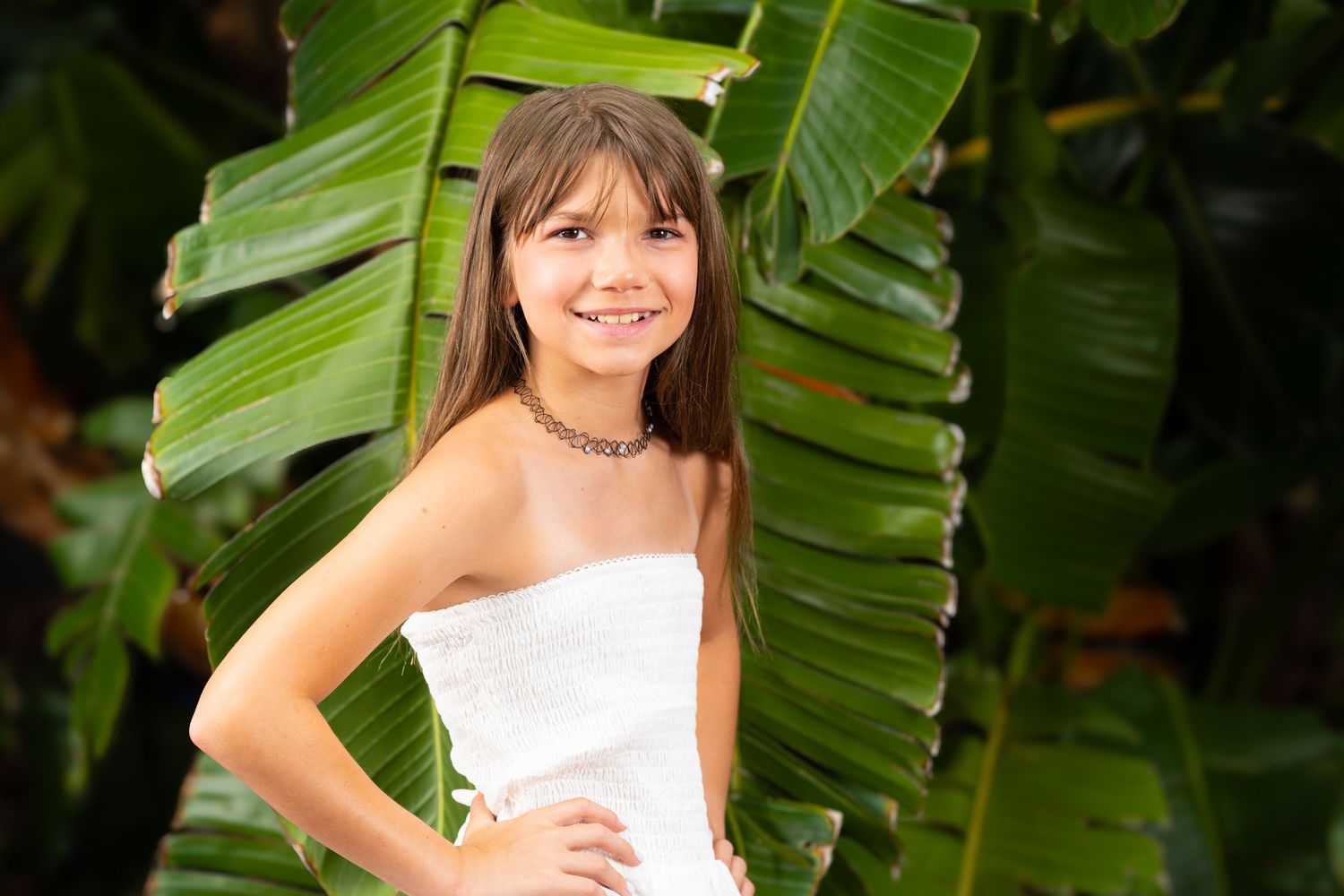 Young girl in white dress smiling outdoors with tropical banana leaves in background.