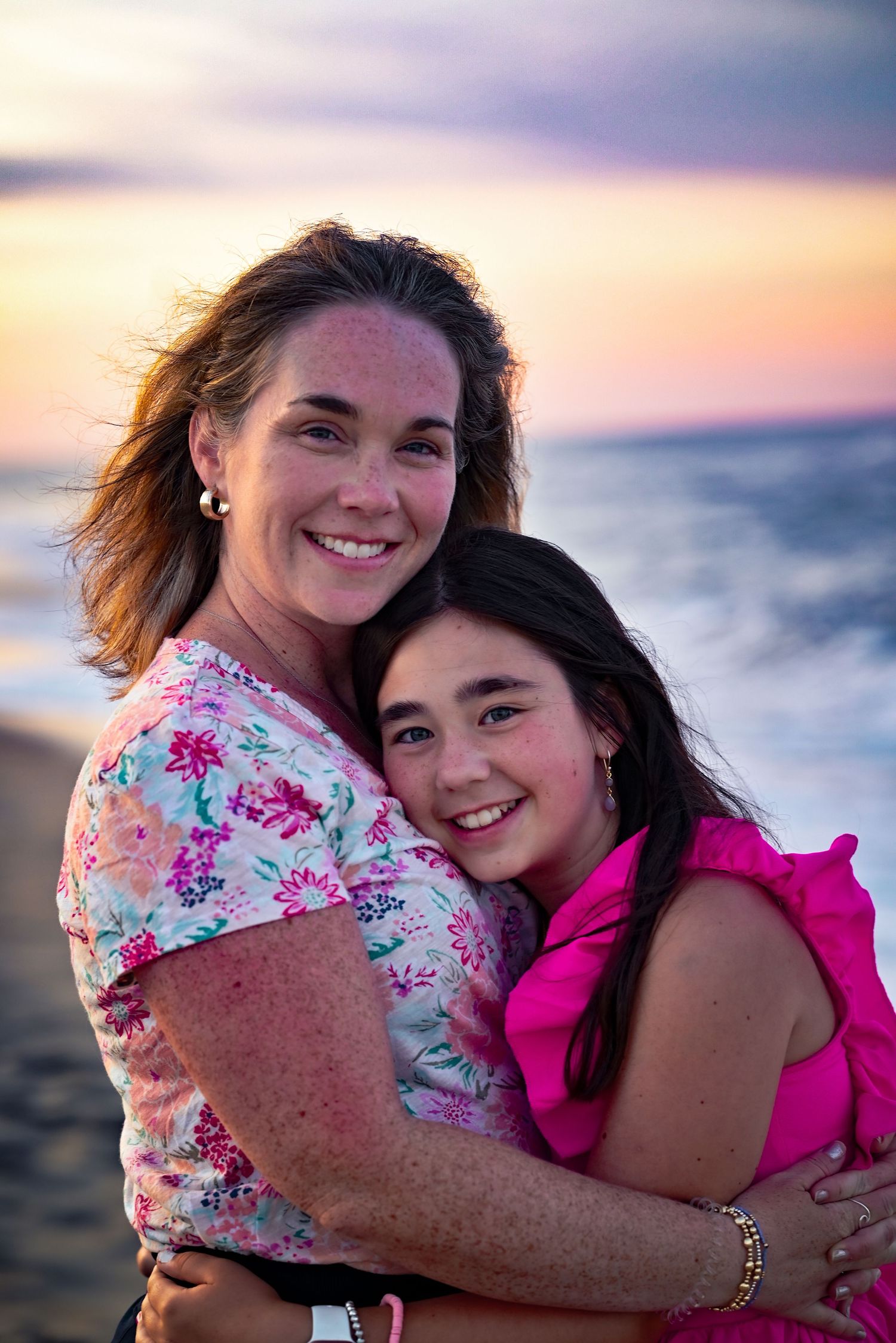 Woman and preteen girl embracing and smiling on a beach at sunset.