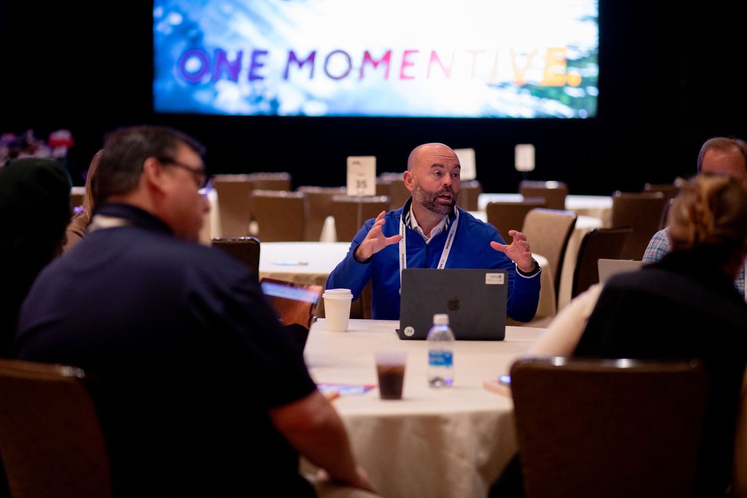 Group talking at a table in front of a screen with branded event graphics at a sales kick off event.