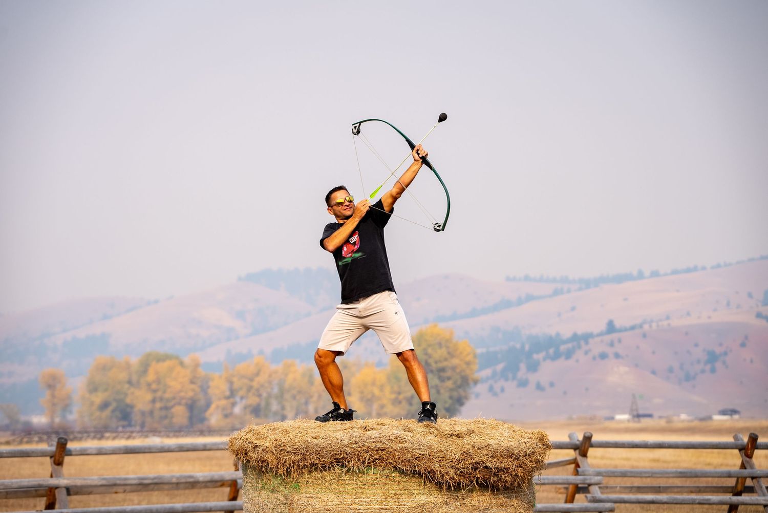 Man standing on on hay bail holding a bow and arrow pointing at the sky.