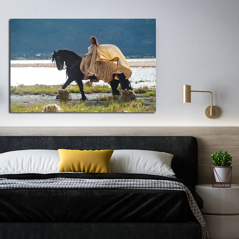 Canvas wall art of a woman in a flowing yellow gown riding a black horse near water, displayed above a modern bed.