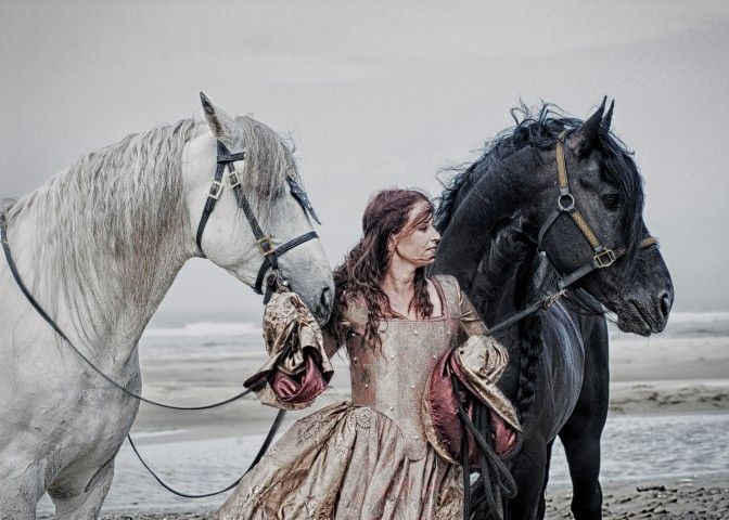 A woman in a vintage gown stands on a beach holding a white and black horse on either side.
