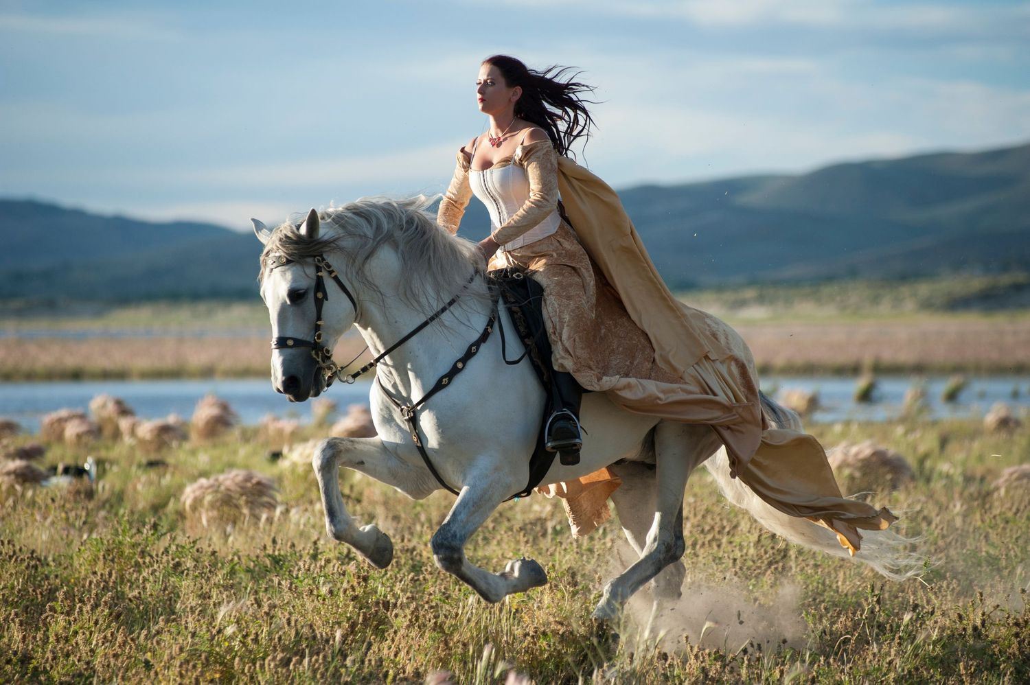Woman in flowing cape rides white horse galloping through scenic landscape with mountains and water in background.