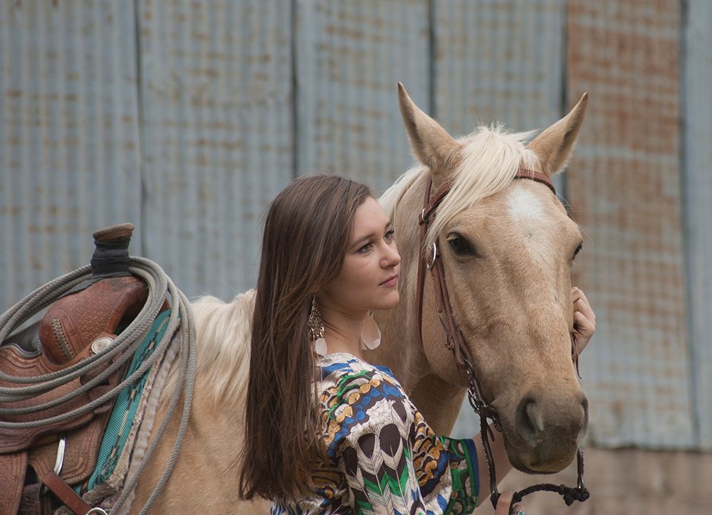 Young woman in floral top affectionately nuzzling a palomino horse with Western saddle outdoors.