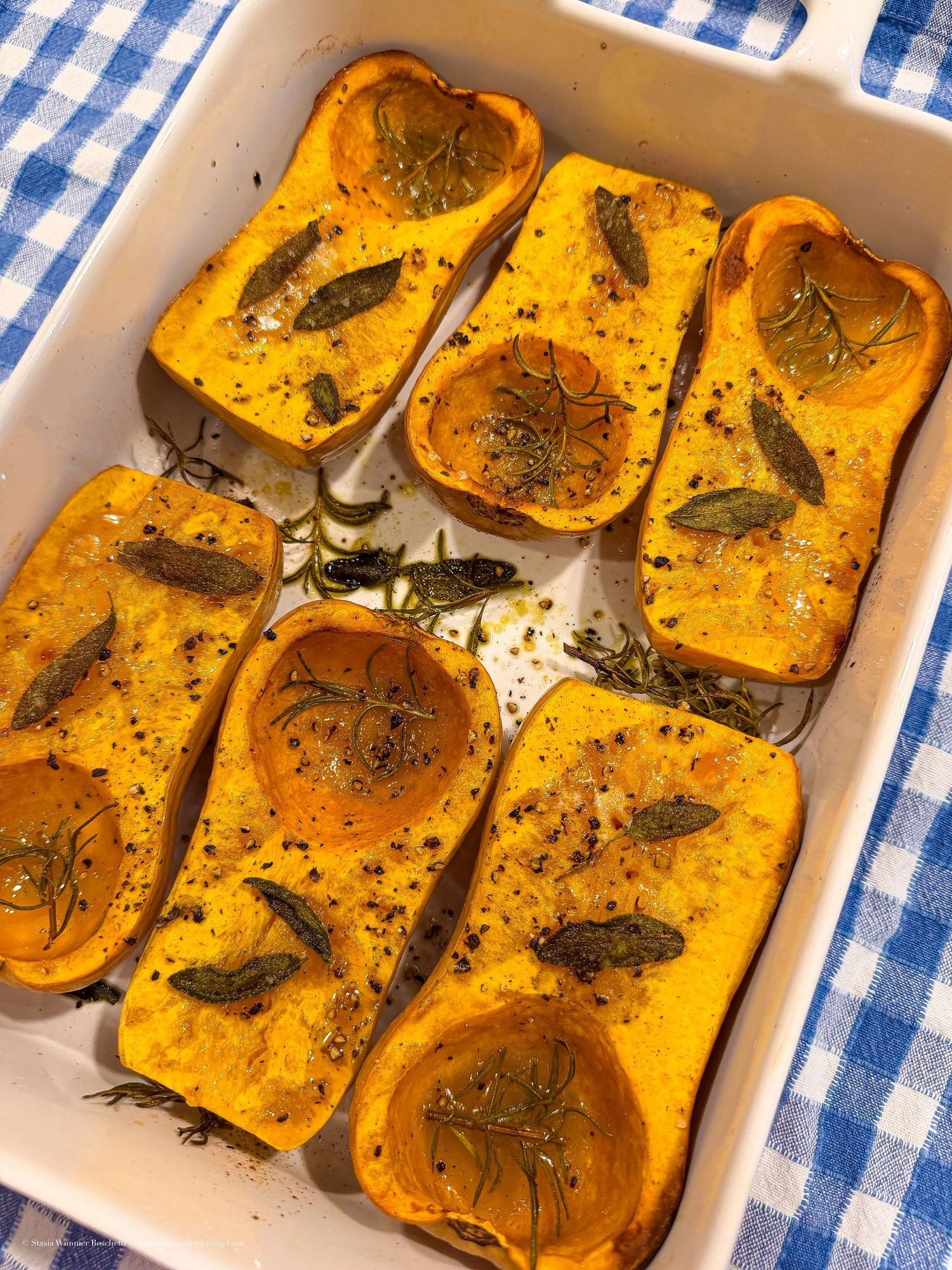 Roasted butternut squash halves with sage and herbs in a white baking dish on a blue checkered tablecloth.
