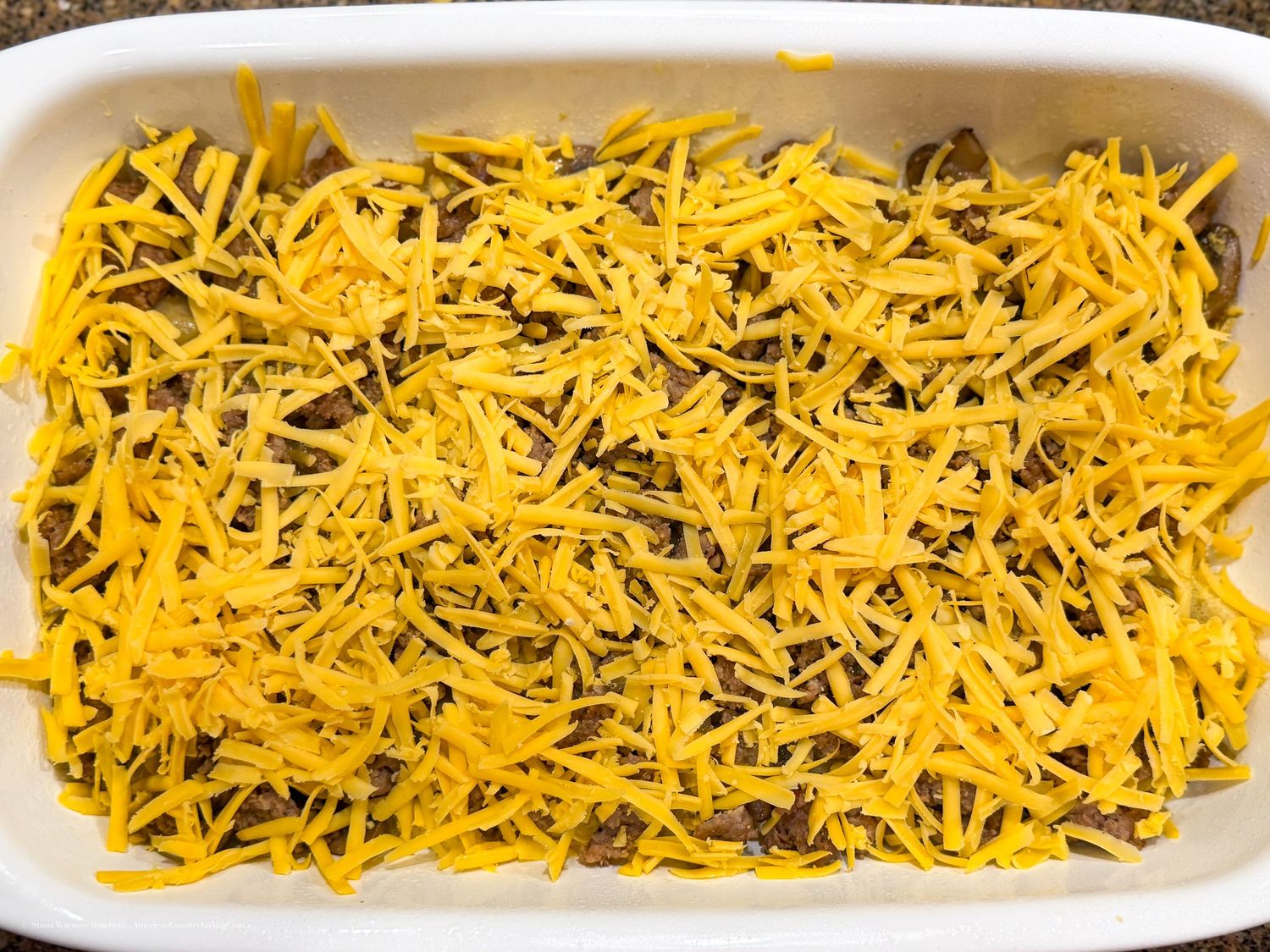 White baking dish topped with shredded cheddar cheese over a meat filling, ready for oven baking.