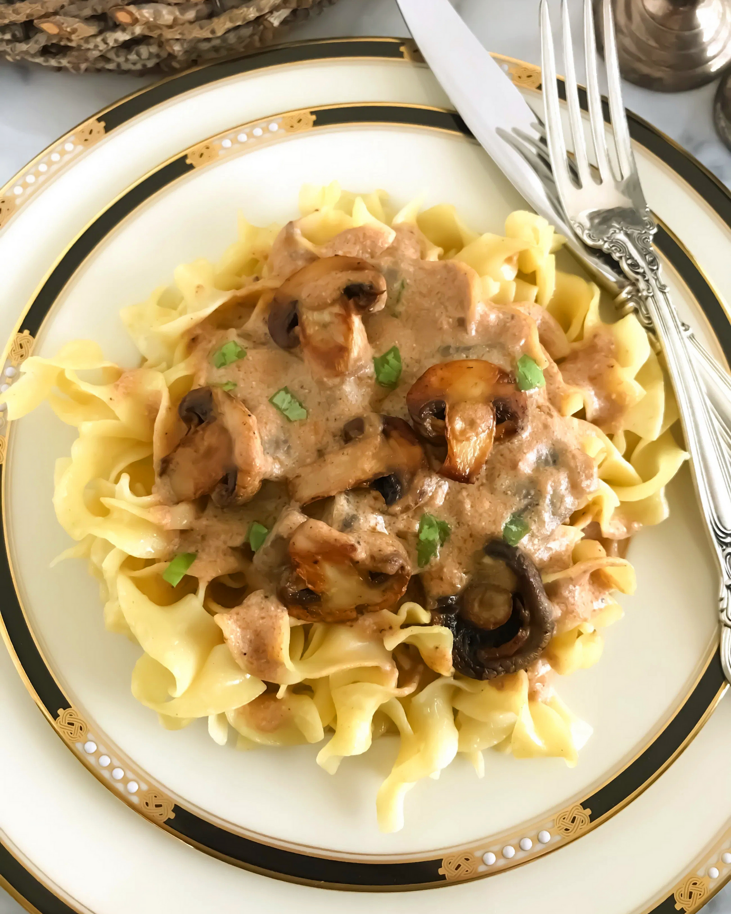 Creamy beef stroganoff with mushrooms served over pasta on a white and gold plate.