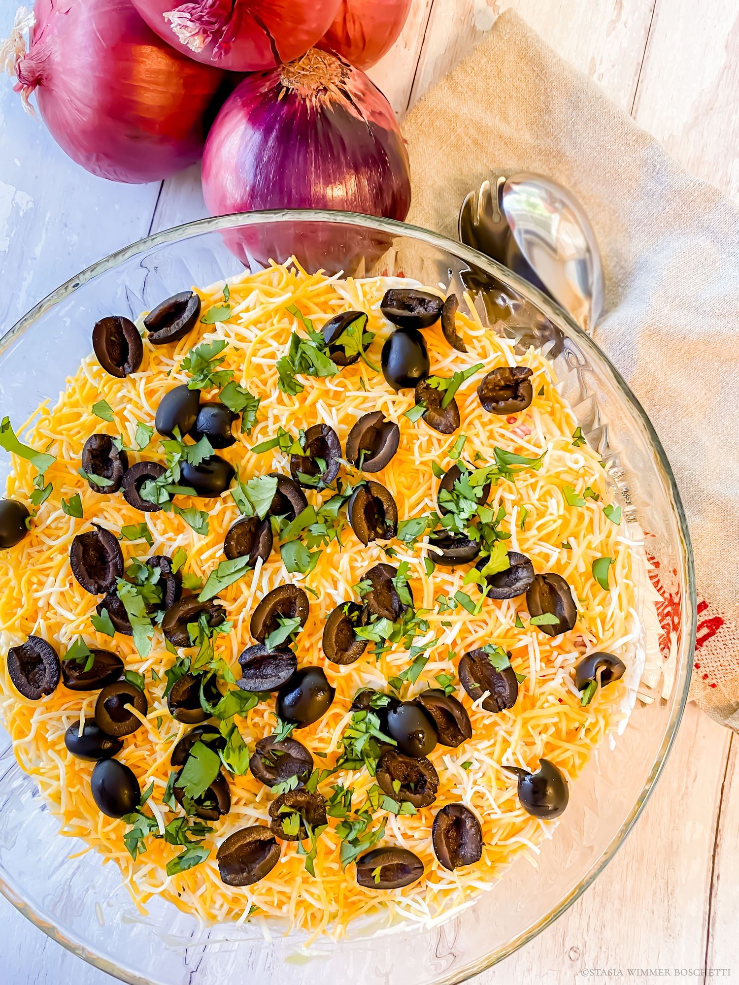 Mexican-style layered dip decorated with black olives, shredded cheese, and herbs on a white wooden surface.