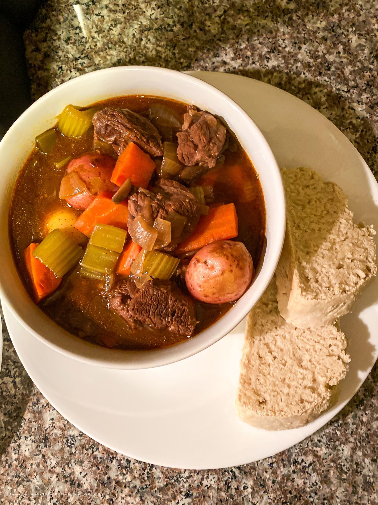 A bowl of beef stew with carrots and celery in rich brown broth sits beside a dusted white plate.