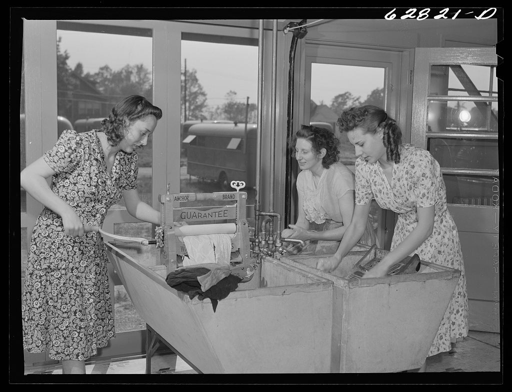 Three women in floral dresses work together at a counter in a 1940s interior setting.