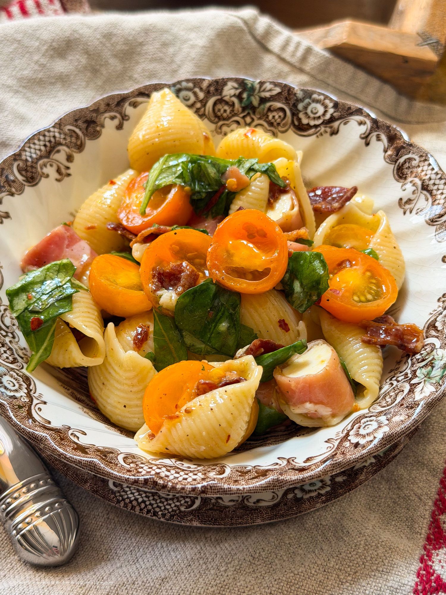 Shell pasta salad with cherry tomatoes, fresh basil, sun-dried tomatoes, and ham in a decorative brown bowl.