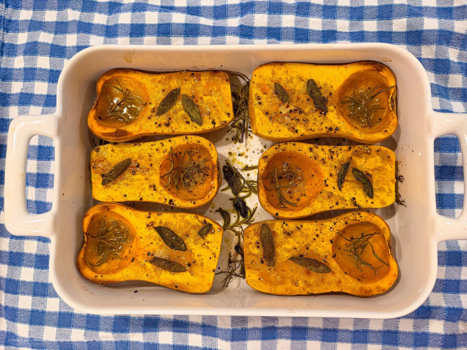 Roasted butternut squash halves with fresh herbs in a white ceramic baking dish on a blue checkered tablecloth.