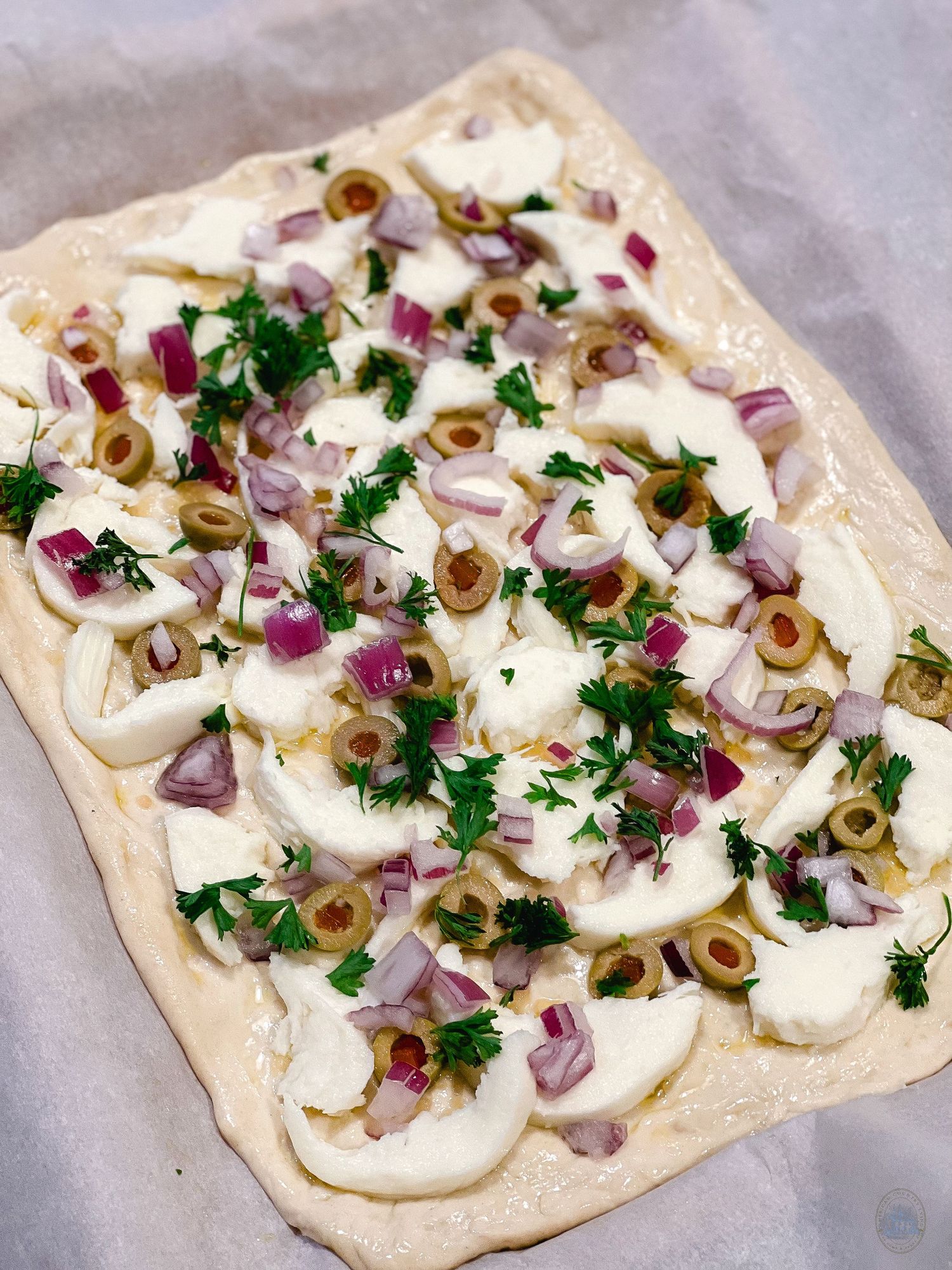 Unbaked flatbread topped with fresh mozzarella, purple onions, green olives and fresh herbs.