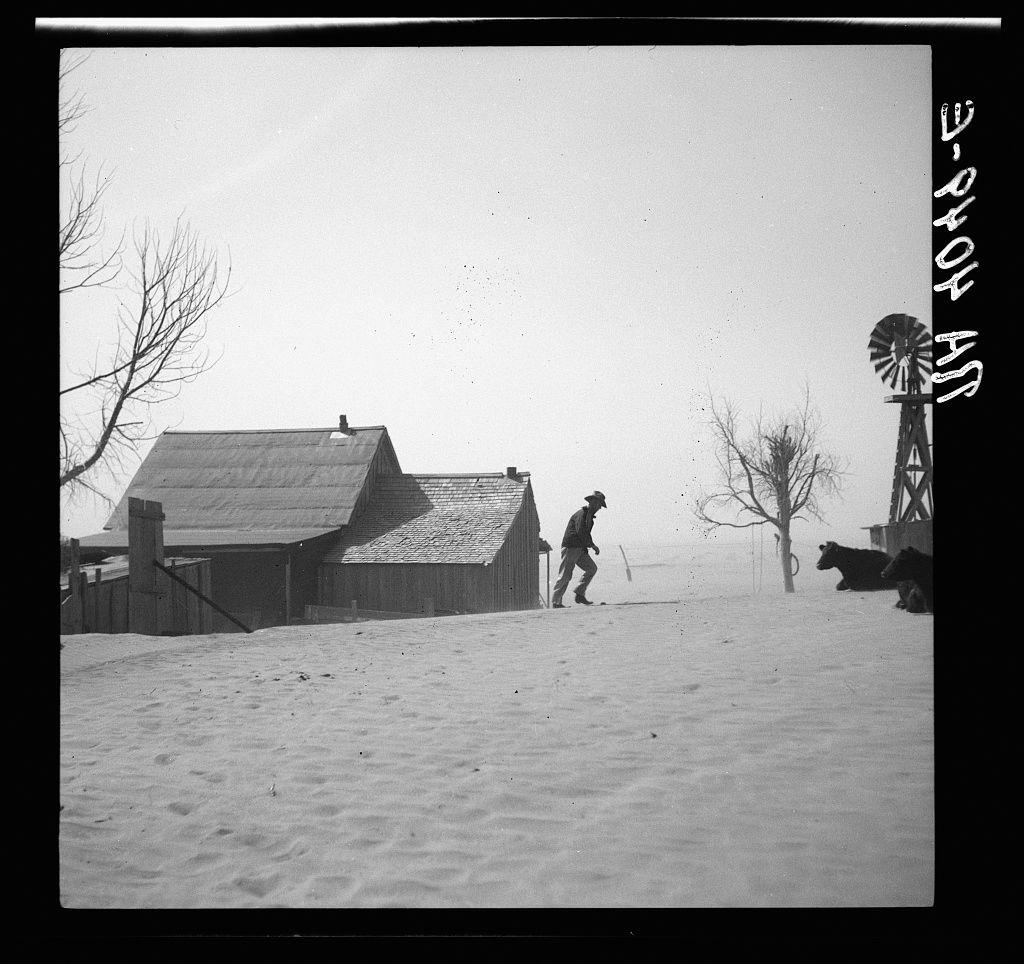 A person climbs up a steep snow-covered slope near a farmhouse in a black and white winter landscape.