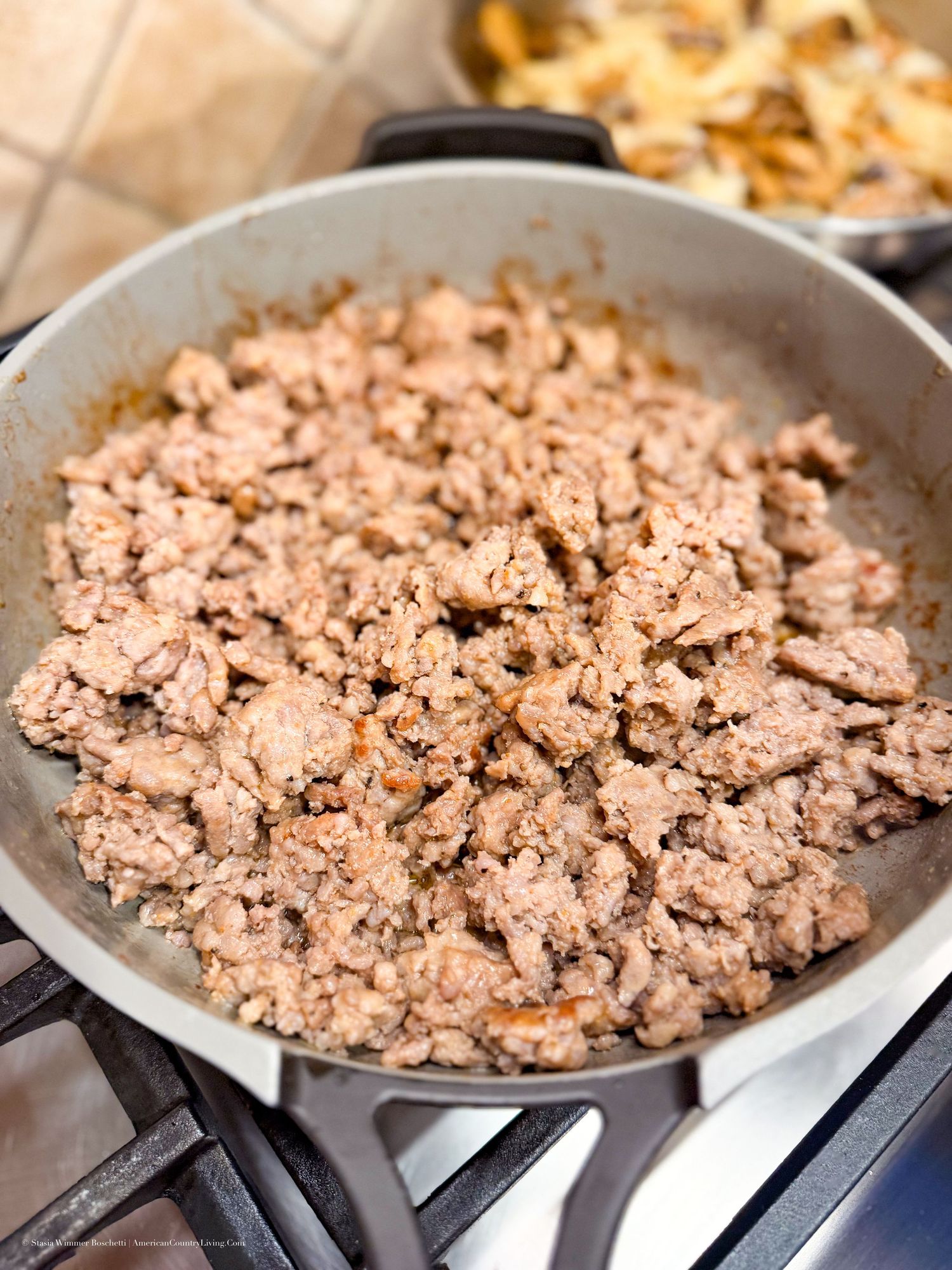 Browned ground meat cooking in a gray skillet on a gas stovetop, seasoned with herbs and spices.