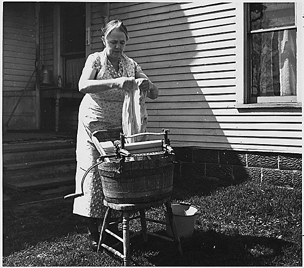 A vintage black and white photograph shows someone doing laundry with an old-fashioned washing machine on a porch.