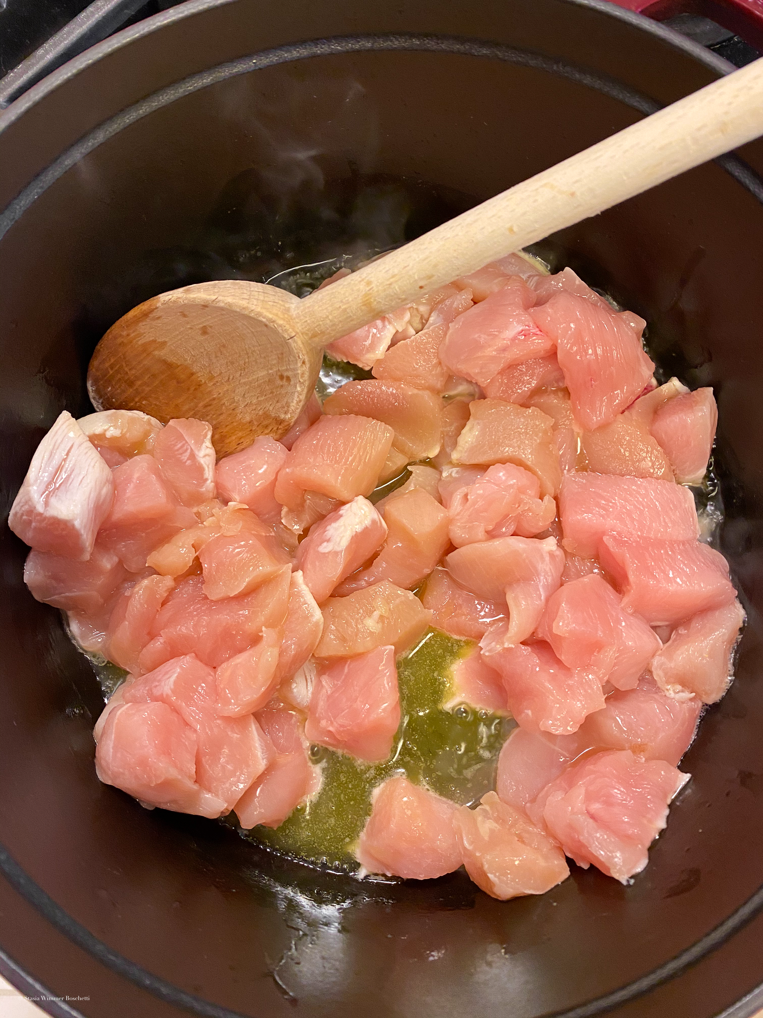 Raw chicken pieces being cooked in a dark pan with wooden utensils.
