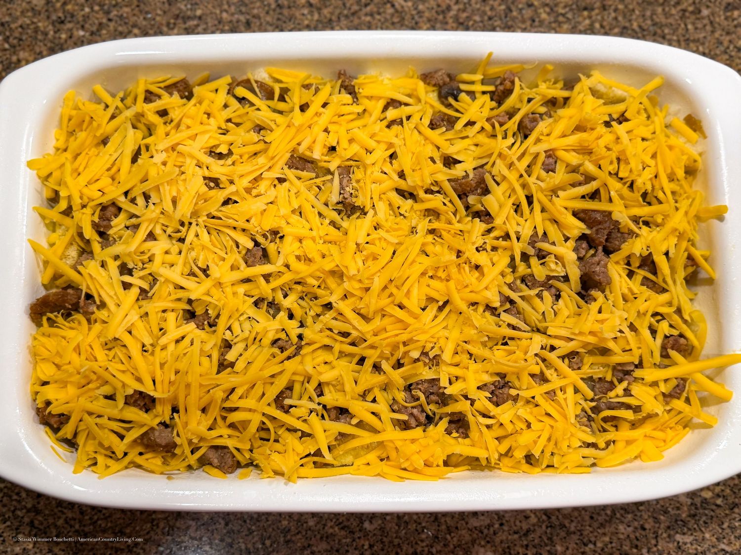 White baking dish filled with seasoned ground beef topped with shredded cheddar cheese, ready to bake.