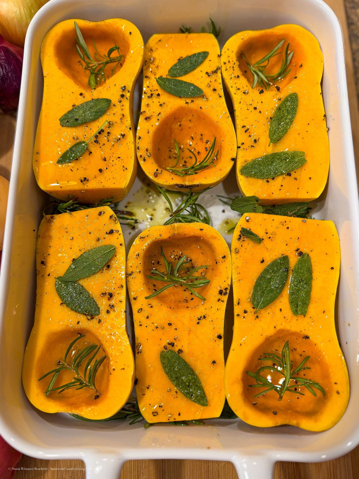 Six halved butternut squash seasoned with herbs, rosemary, and sage in a white baking dish, ready to roast.