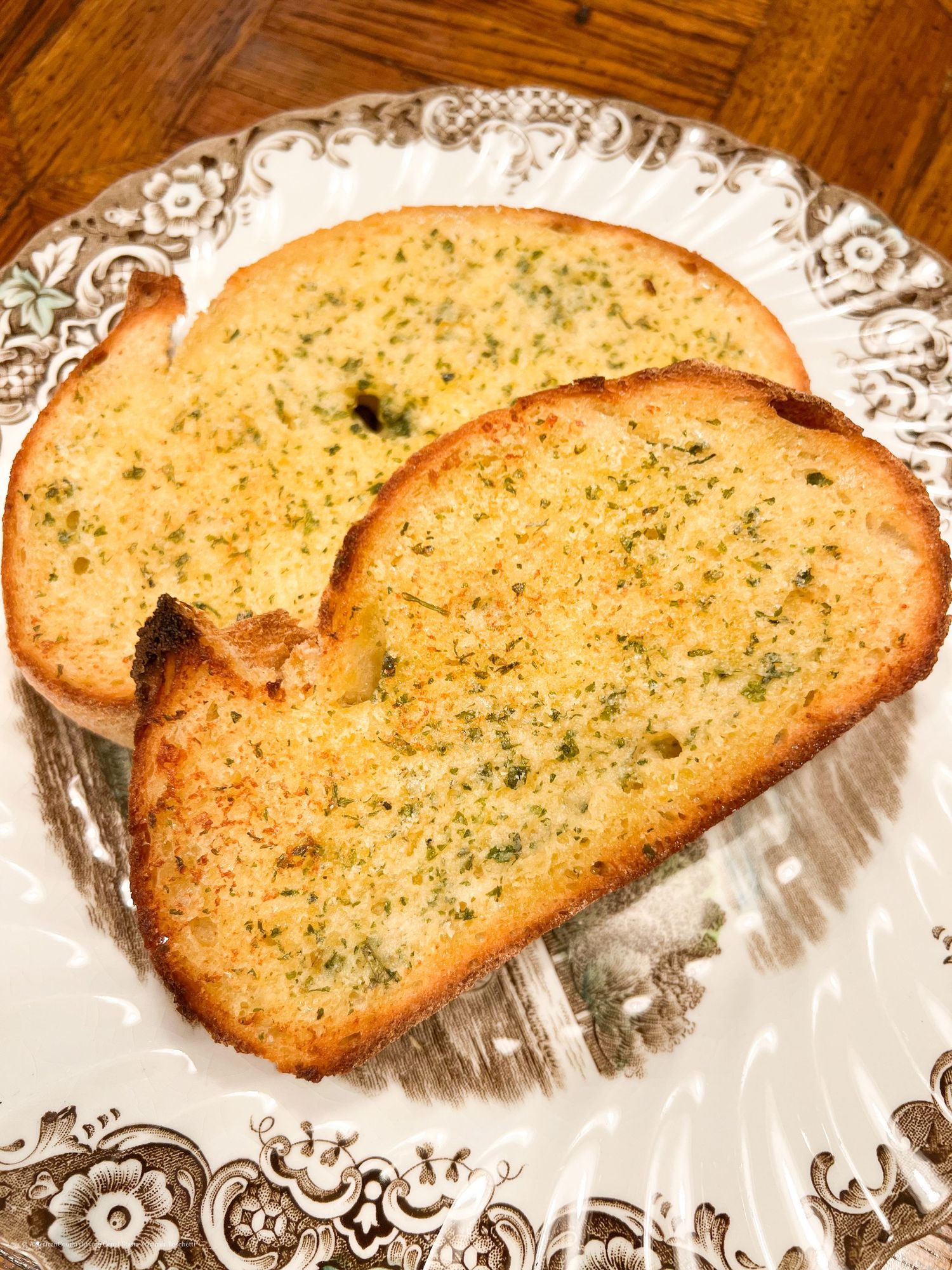 Slices of golden garlic bread with herbs arranged on a decorative white plate with ornate pattern.