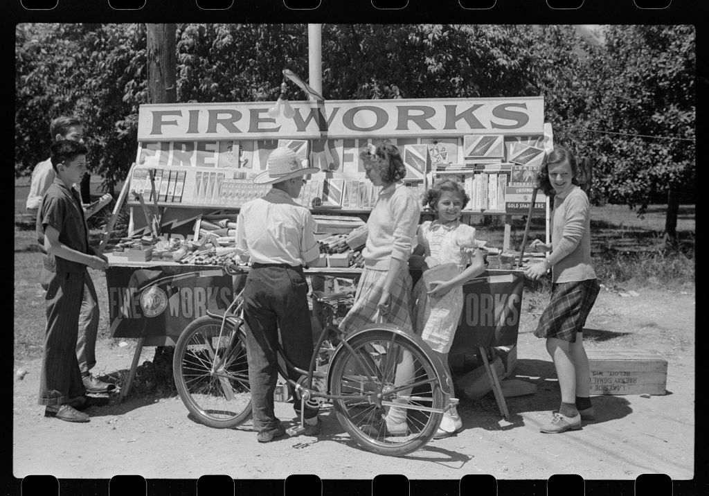 A vintage black and white photograph of a fireworks stand with customers gathered around bicycles in the 1930s.