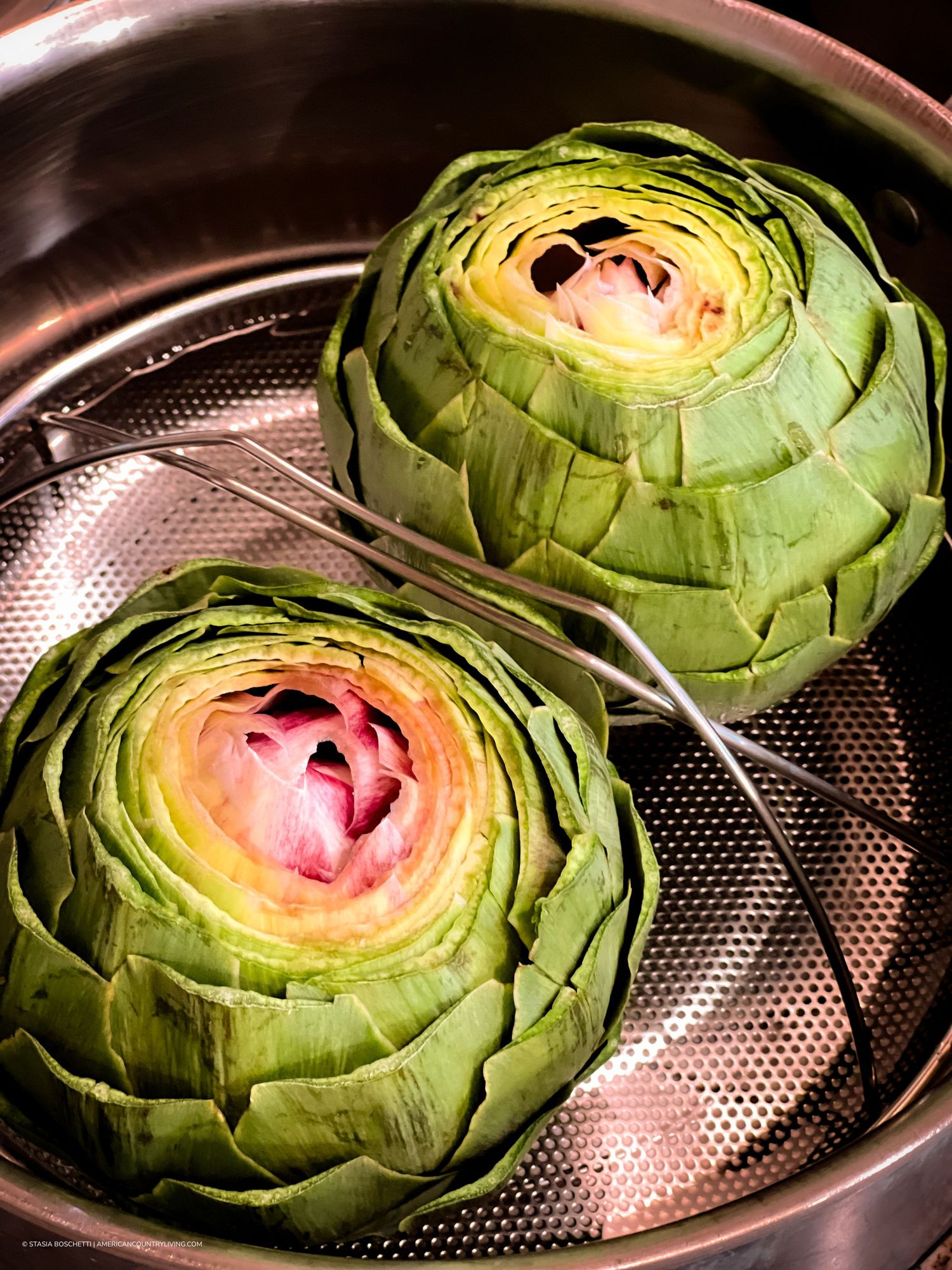 Two fresh artichokes steaming in a stainless steel steamer basket, showing vibrant green leaves.
