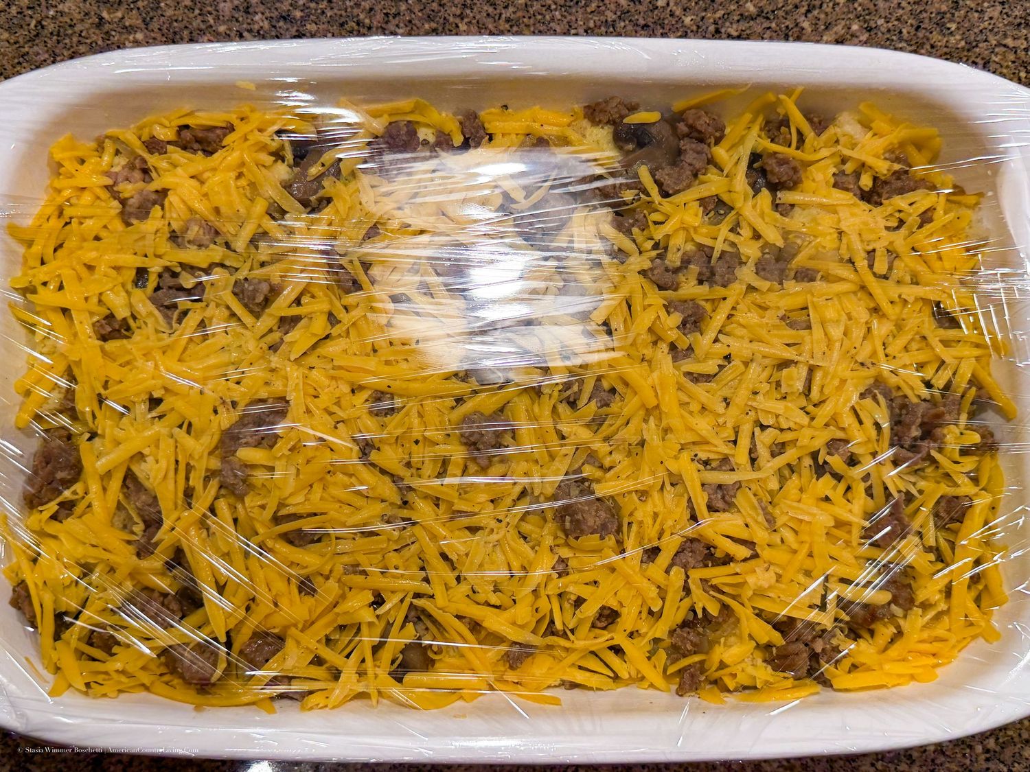 Shredded cheddar cheese topping a casserole dish with ground beef, covered in plastic wrap, ready to bake.