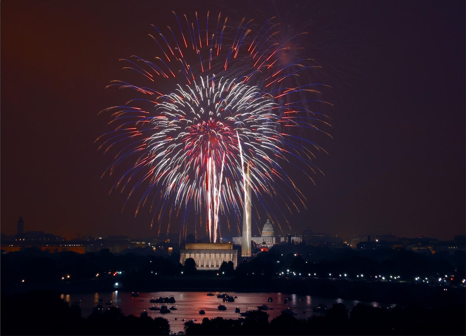Colorful red and white fireworks burst over Washington DC monuments and reflecting pool at night during celebration.