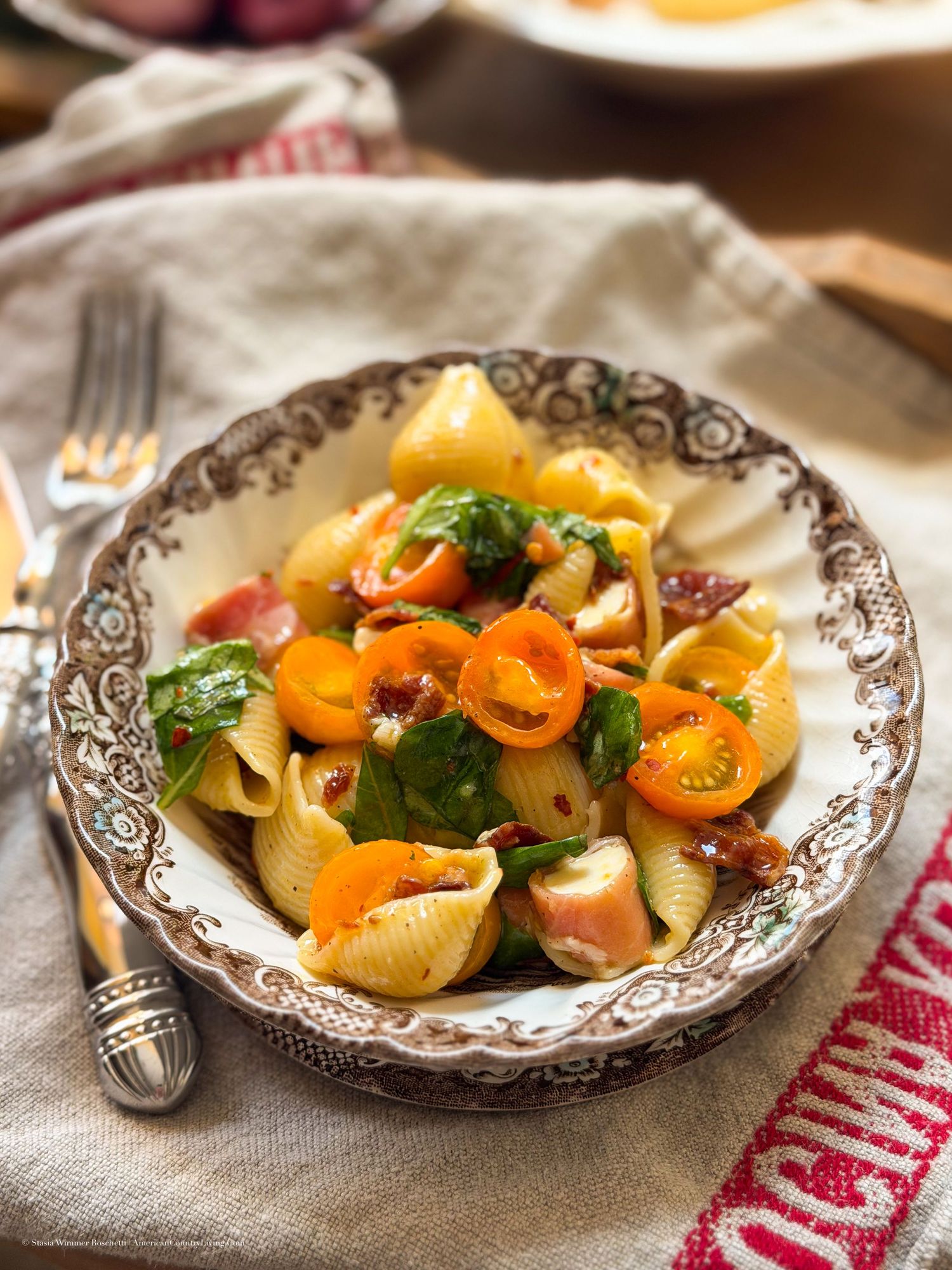 Shell pasta salad with cherry tomatoes, bacon, and fresh basil served in a decorative bowl on a rustic table.