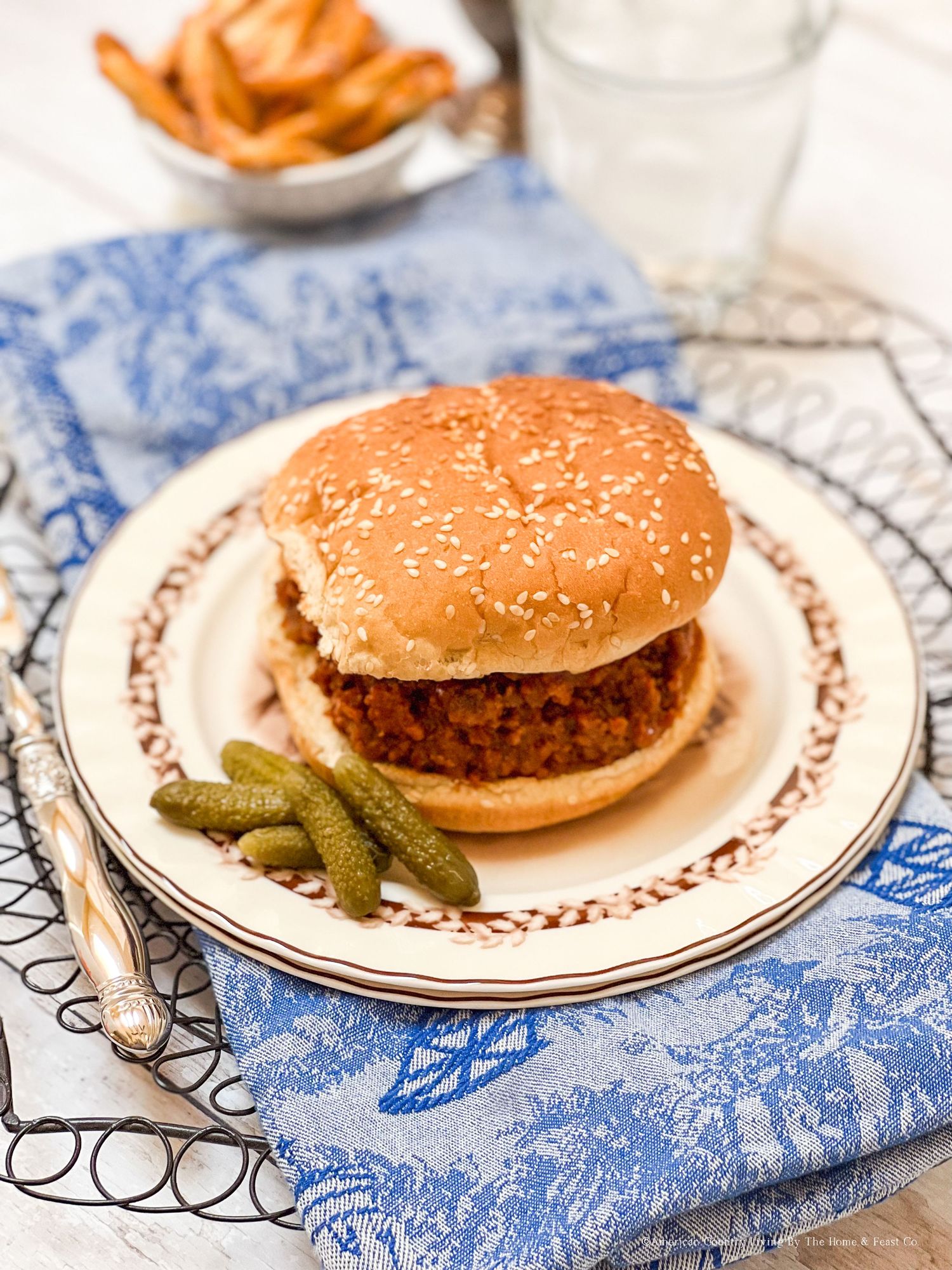Sloppy joe sandwich on toasted bun plated with pickles on blue and white patterned tablecloth.
