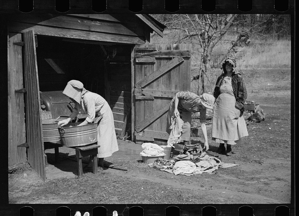 Vintage black and white photograph showing people doing laundry outdoors near a wooden structure in a rural setting.