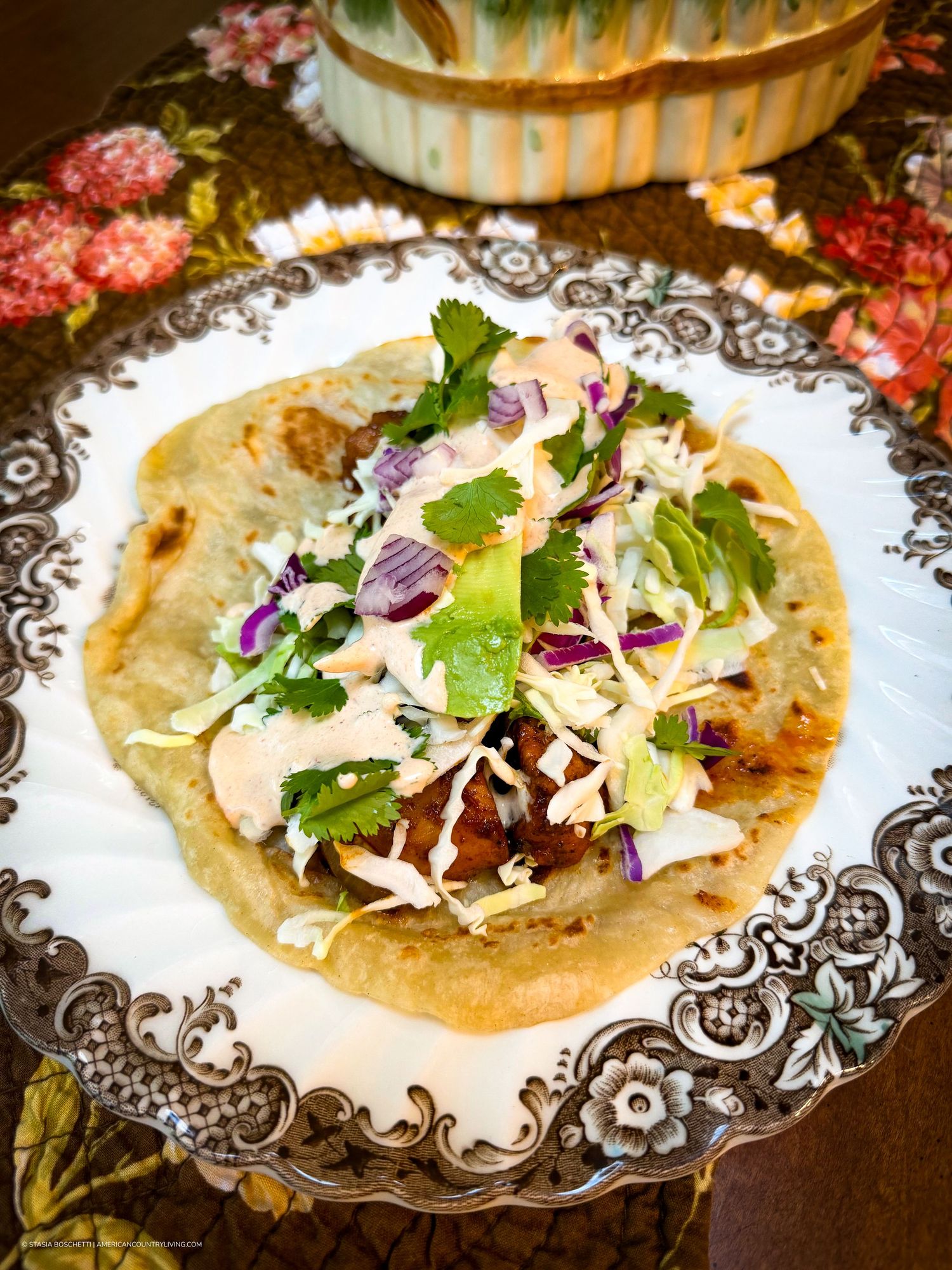 A gourmet fish taco on a flour tortilla topped with coleslaw, cilantro, red onion, and chipotle cream sauce on an ornate plate.