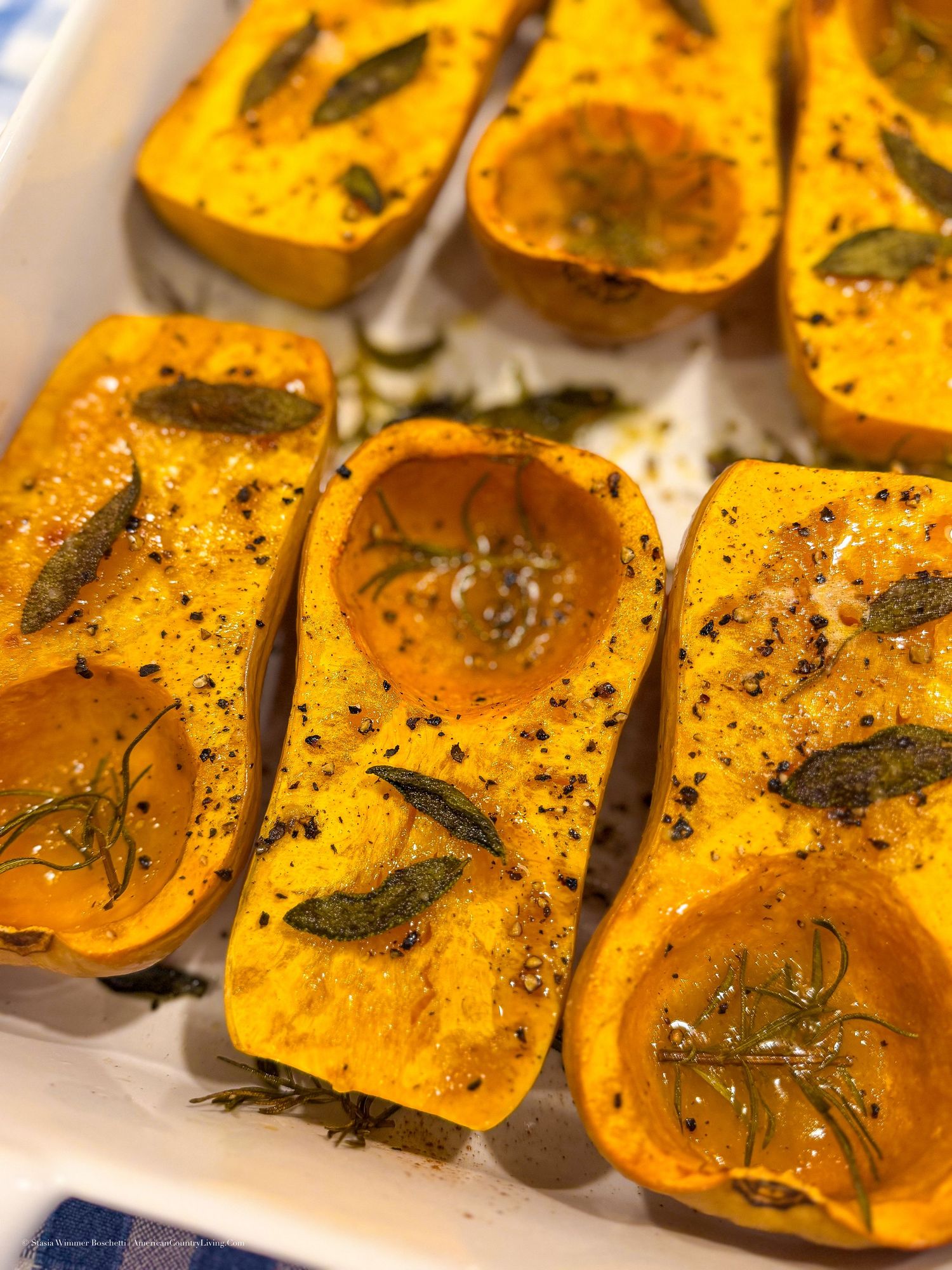 Roasted butternut squash halves seasoned with herbs, sage, rosemary, and black pepper in a white baking dish.