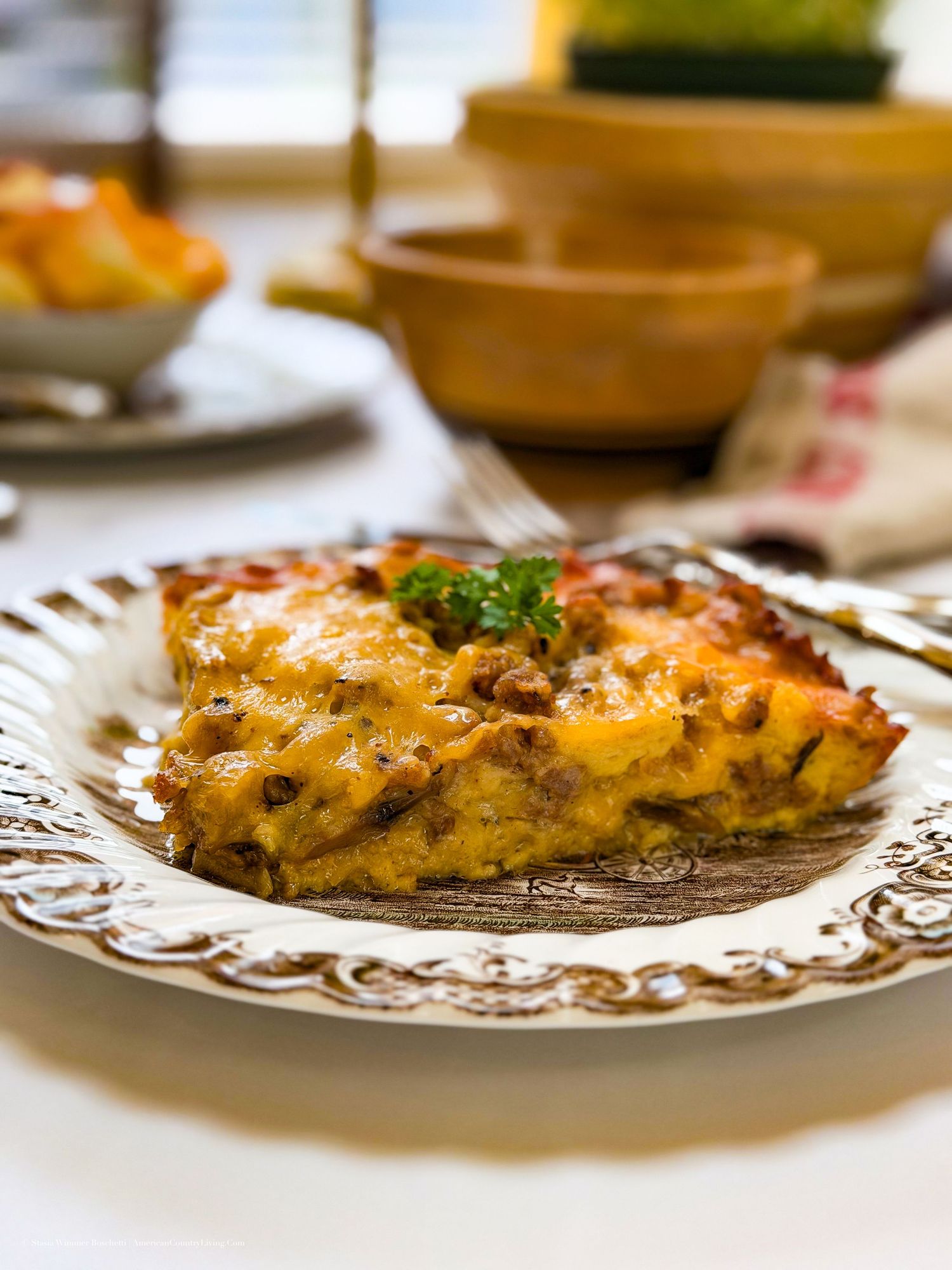 A golden cheesy casserole slice with herbs on a decorative white plate, served on a rustic dining table.