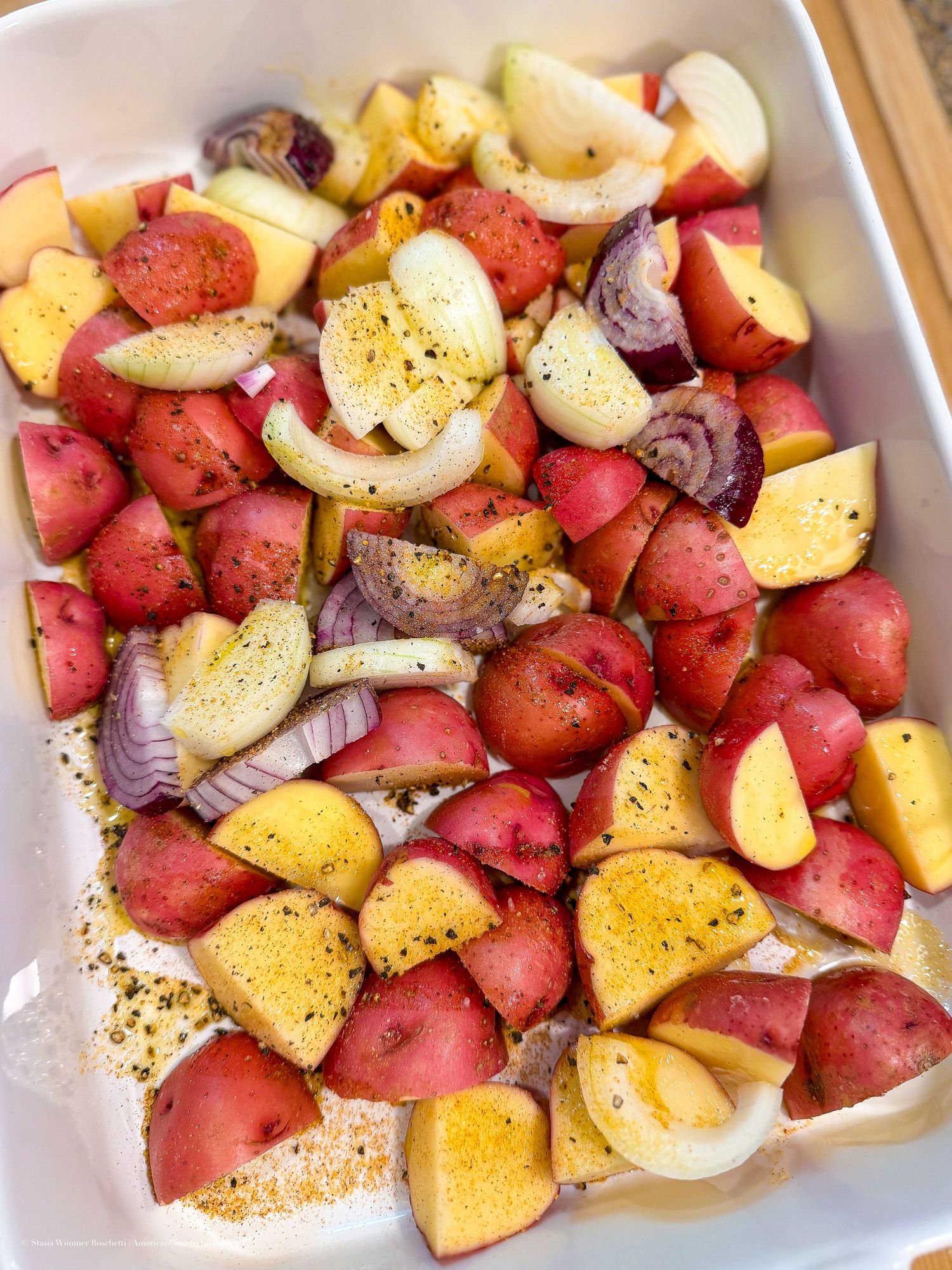 Red potatoes and onions seasoned with pepper in a white baking dish, ready for roasting.