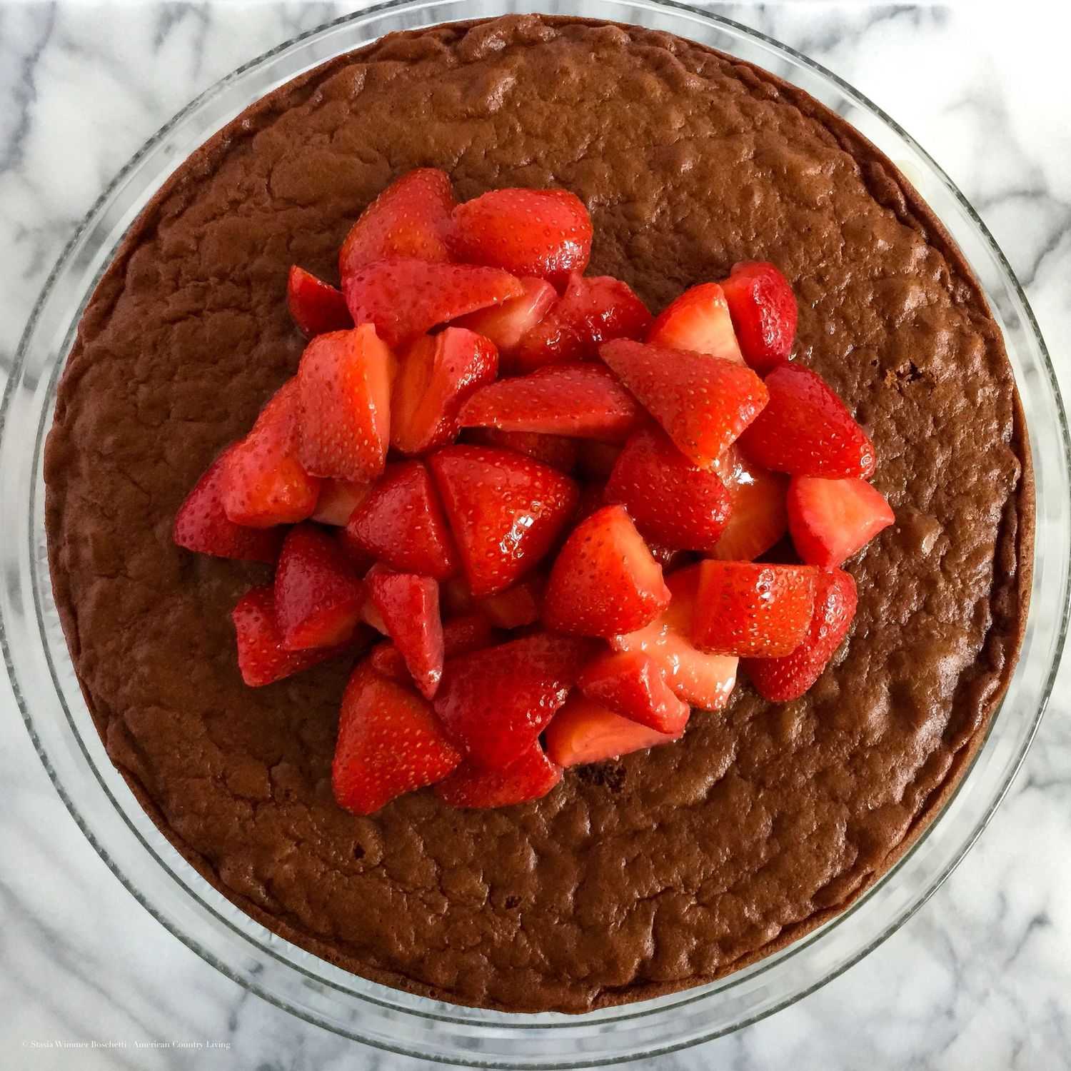Chocolate brownie cake topped with fresh sliced strawberries in a glass dish on marble surface.