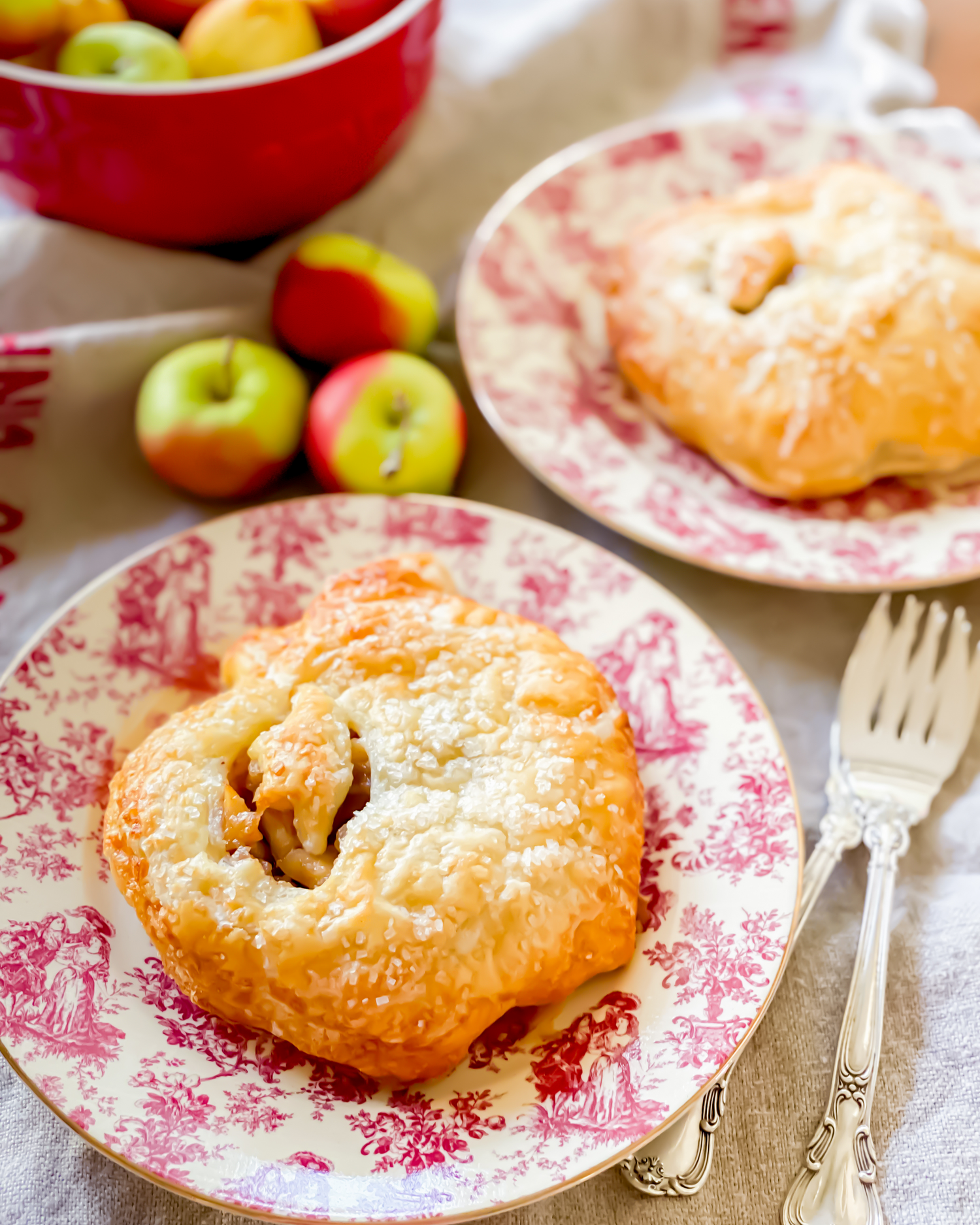 Golden apple-shaped pastries served on vintage pink floral plates with fresh apples in a red bowl.