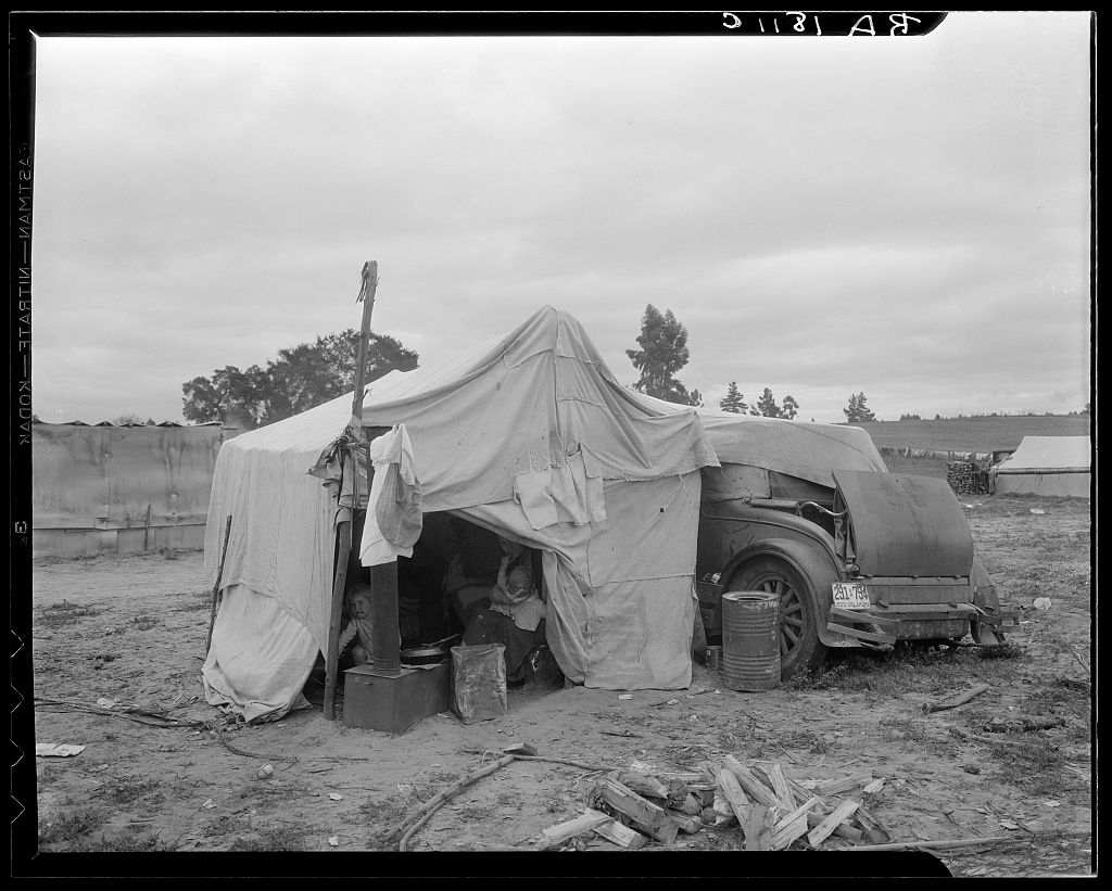 A black and white photograph shows several makeshift tents and temporary shelters set up in a rural field during the 1930s.