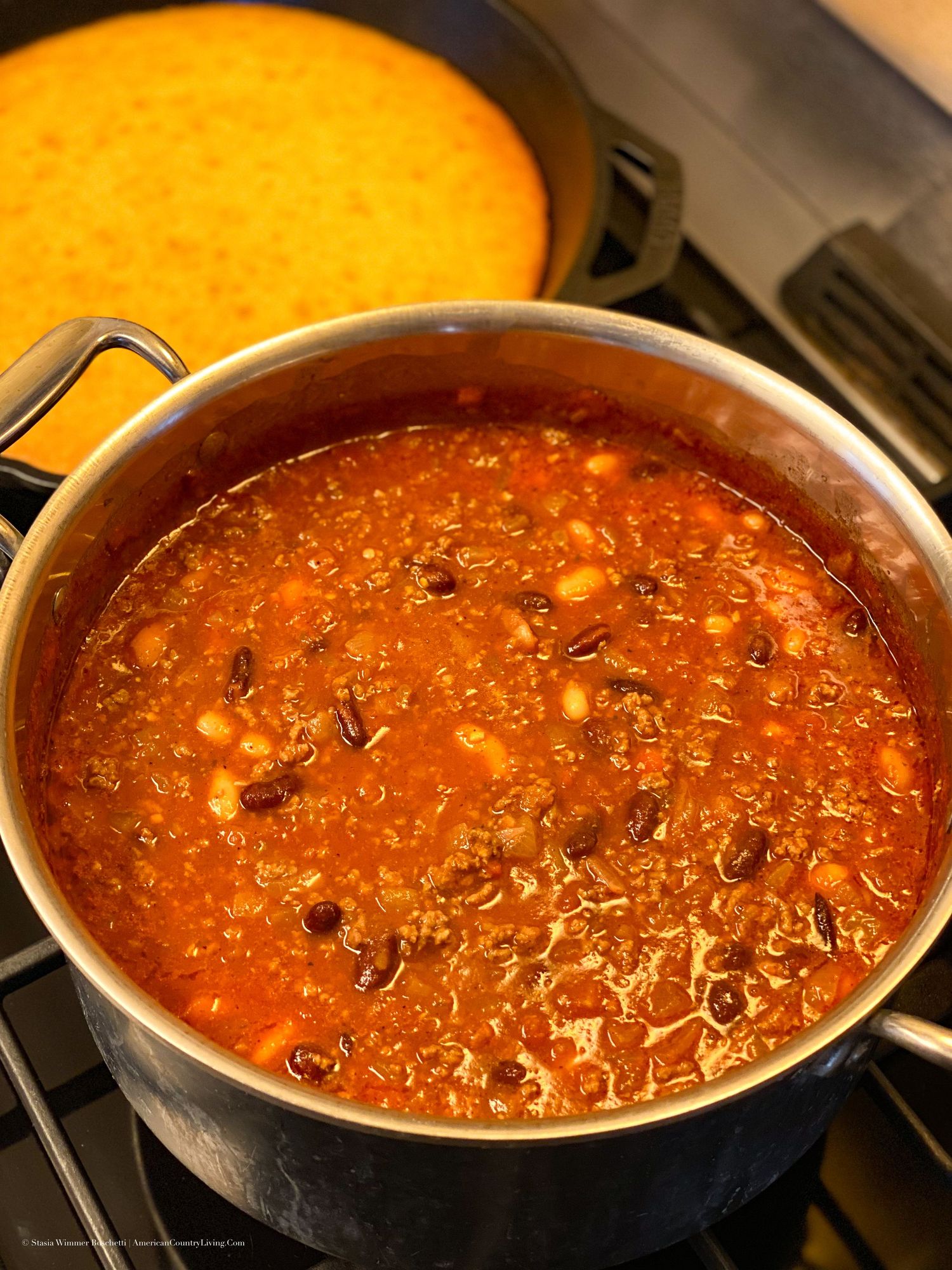 A pot of hearty chili with beans simmering on a stovetop, with golden cornbread visible in a skillet behind it.