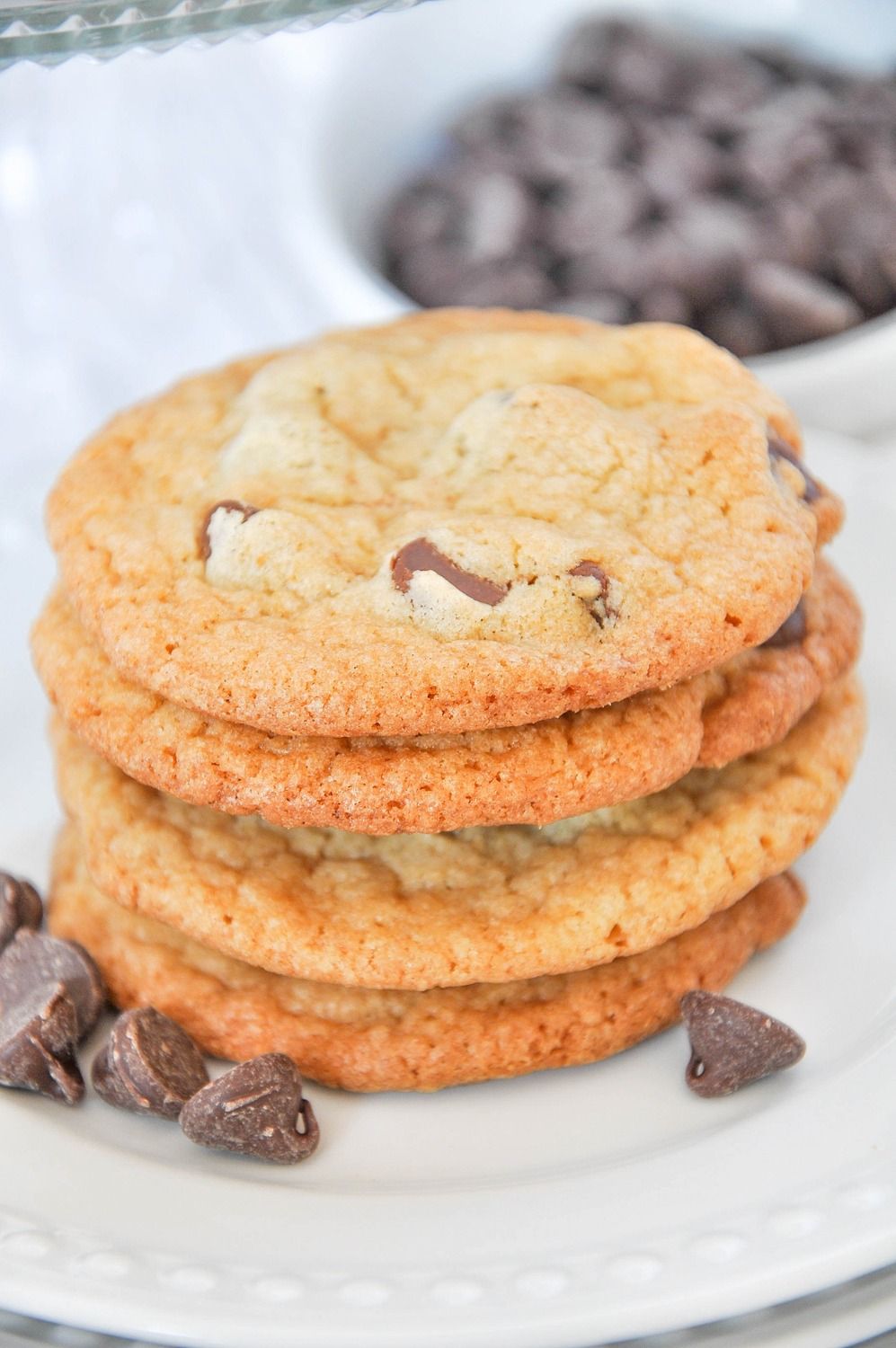 Stack of golden chocolate chip cookies on a white plate with chocolate chips scattered around and a bowl in background.