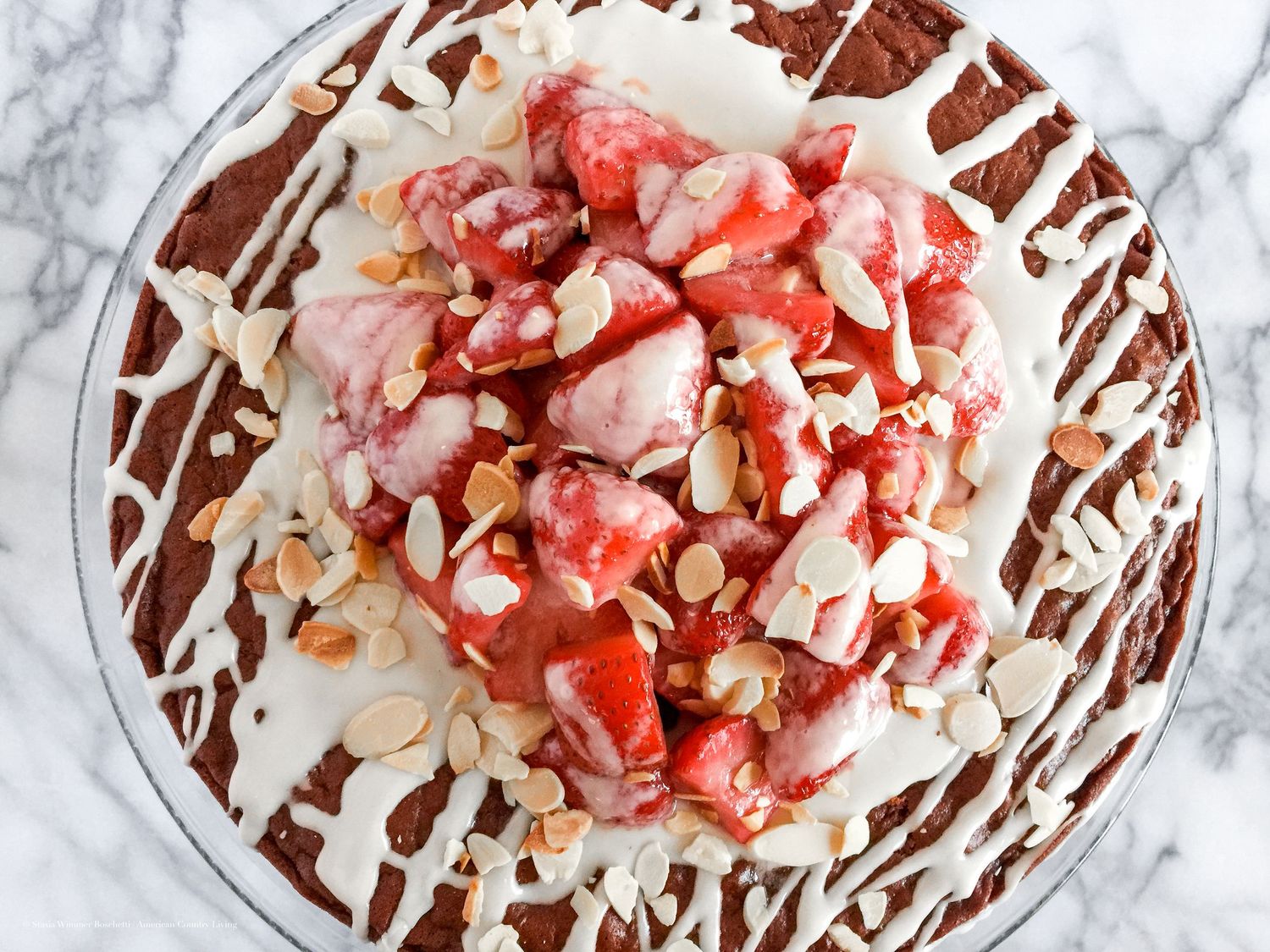 Chocolate layer cake with white glaze, fresh strawberries, and toasted almonds on a glass cake stand.