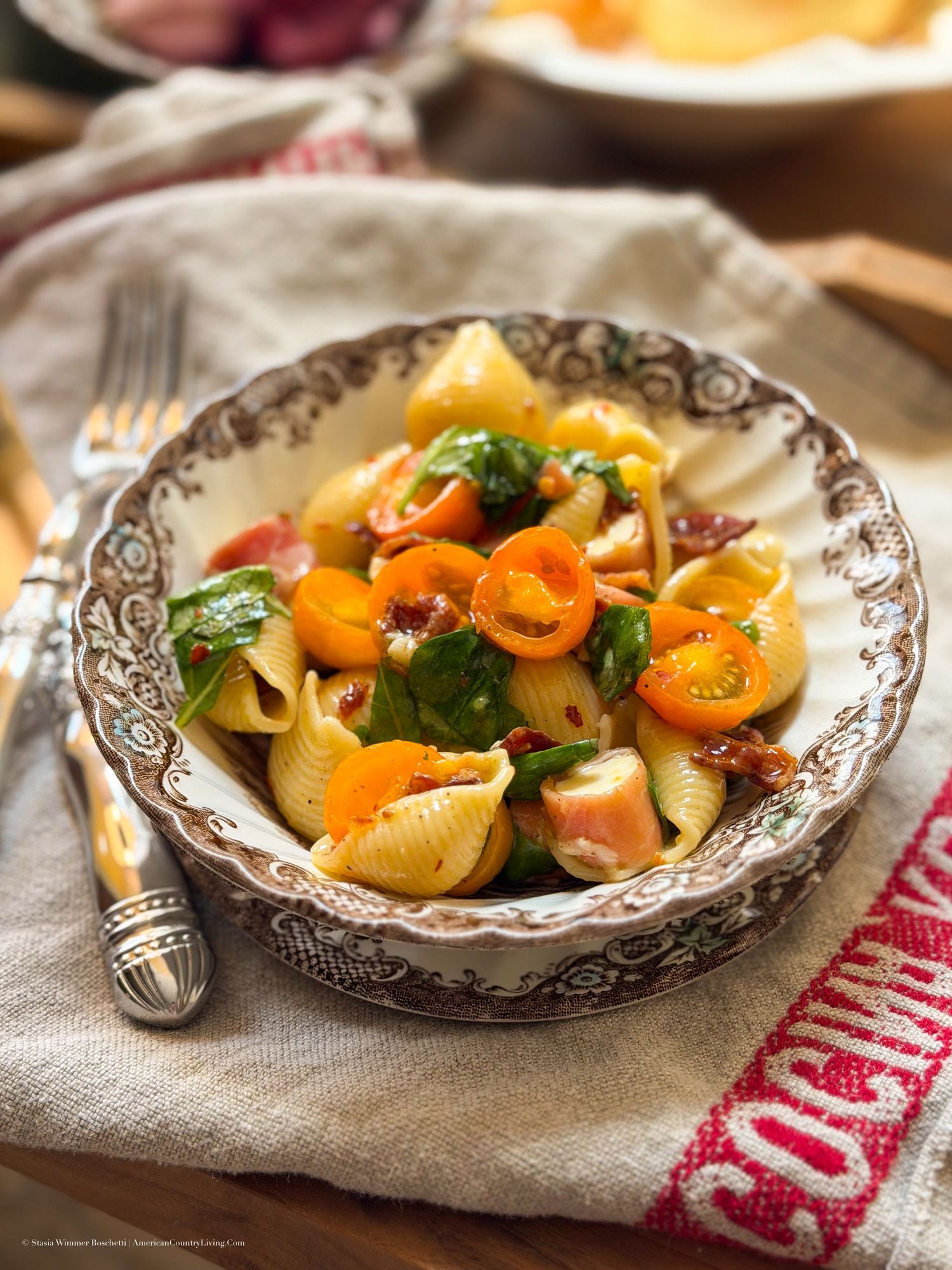 A decorative bowl filled with shell pasta salad, cherry tomatoes, spinach, and sun-dried tomatoes on a rustic table.