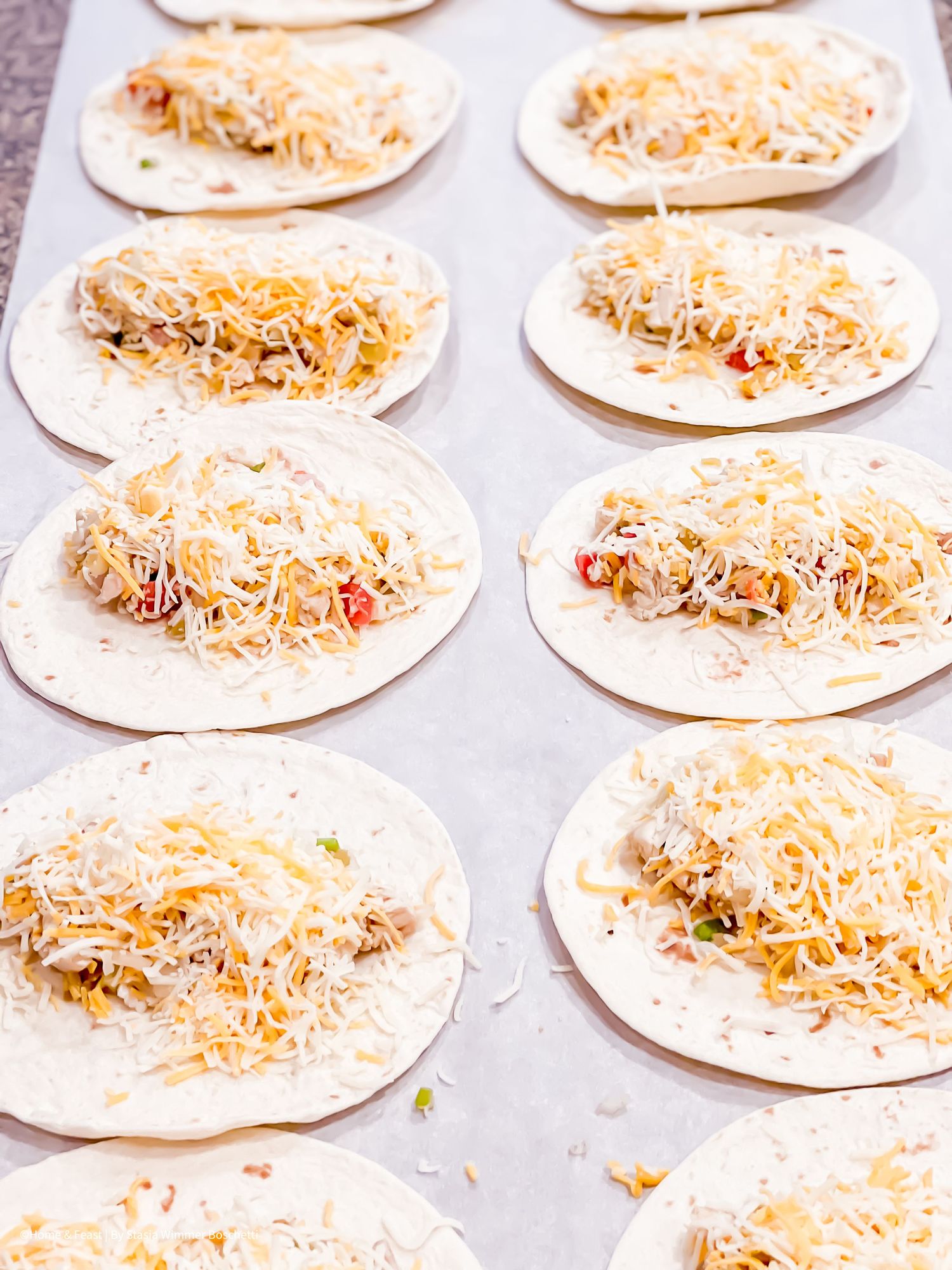 Tortillas topped with shredded cheese and ingredients arranged in rows on a baking sheet.